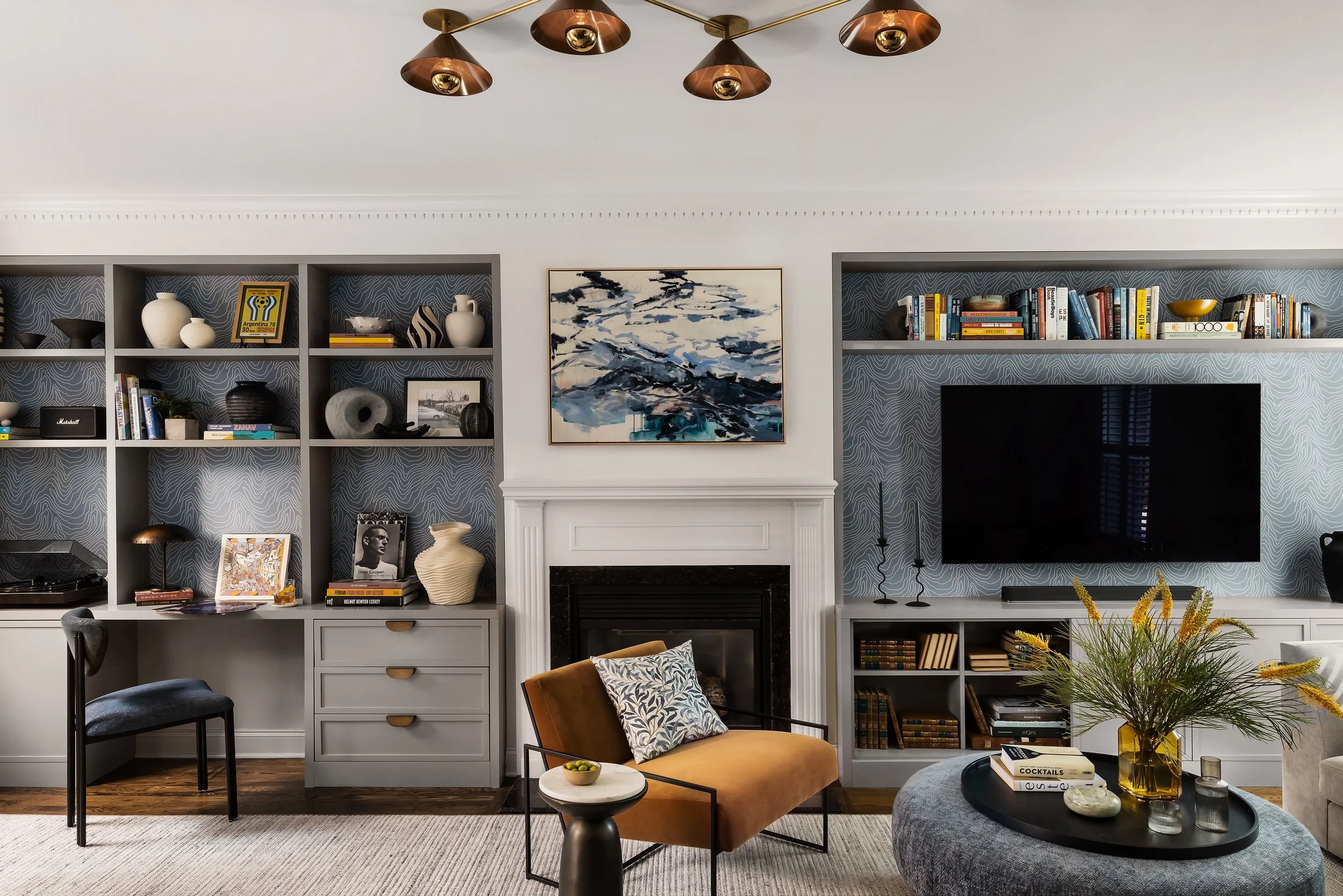 Living room in a South End brownstone with custom gray built-ins on either side of a marble fireplace, patterned wallpaper backing the shelves, a brass multi-arm pendant, and a mustard velvet chair.