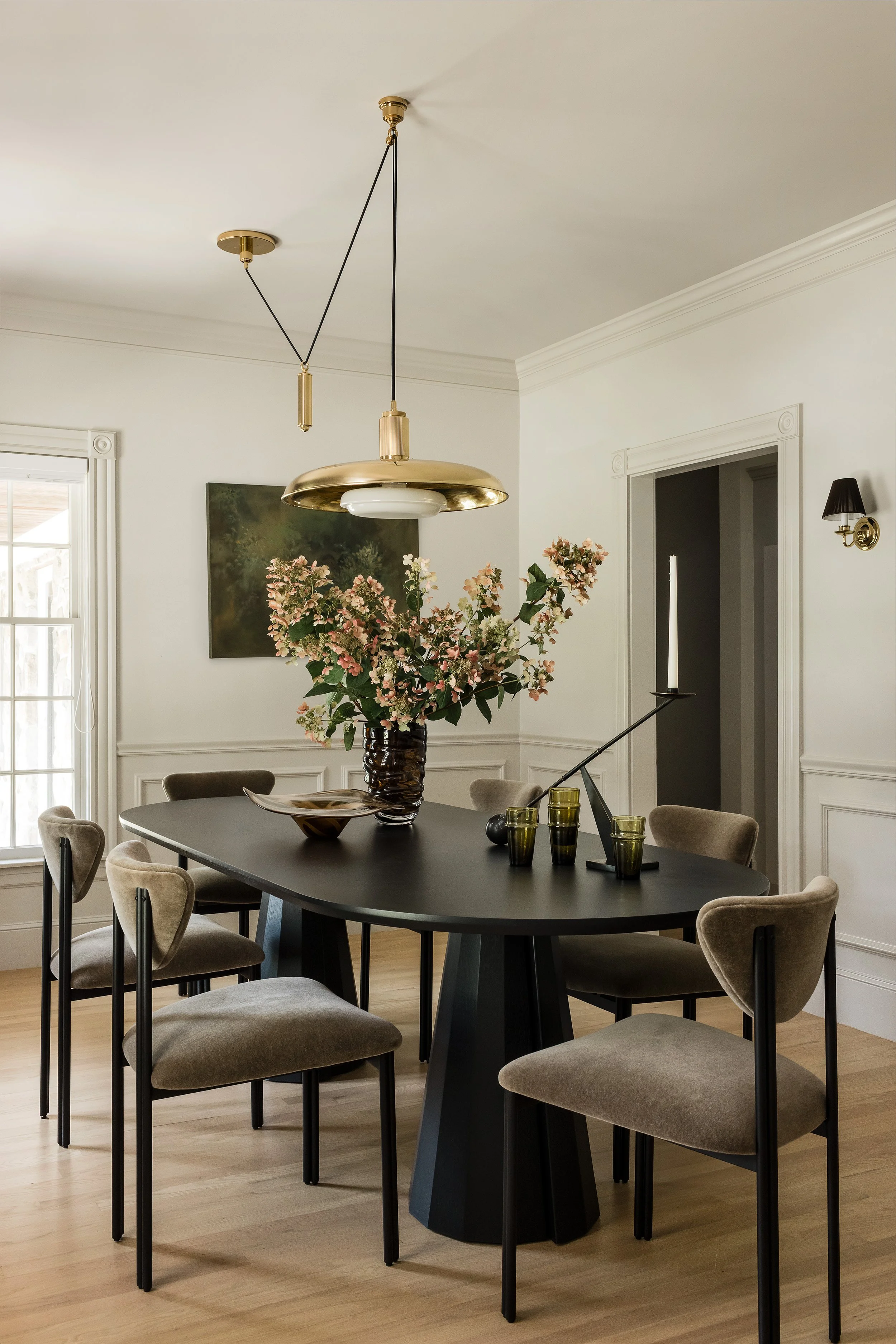 Wide view of the dining room with six upholstered chairs around a black oval table, a large brass disc pendant, and hydrangeas in a smoked glass vase.