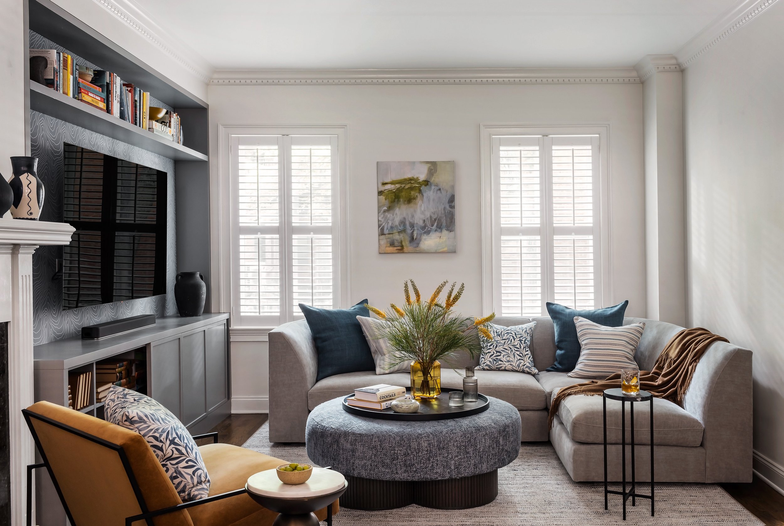 Wide view of a South End living room with a gray sectional sofa, patterned throw pillows, white plantation shutters, and a media console opposite the fireplace.
