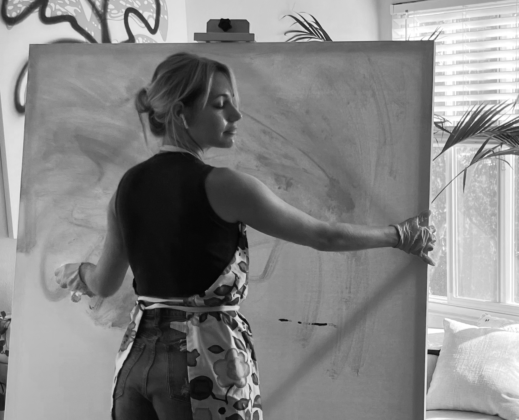 A woman with air pods, wearing a sleeveless top and printed apron, is painting on a large canvas in a well-lit room with a window, plants, and pillows.