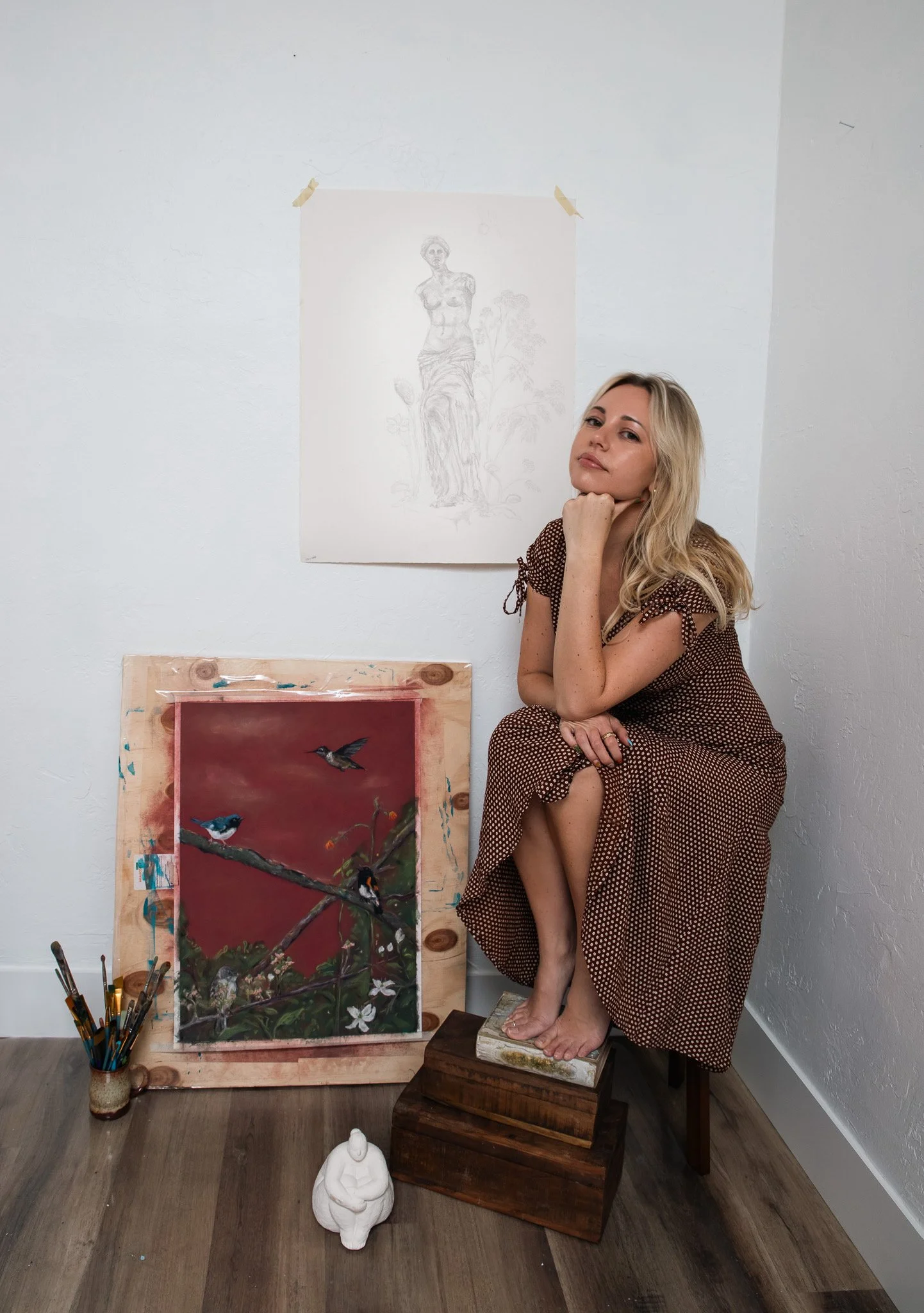 A woman with blonde hair in a brown polka dot dress is sitting on a stack of wooden boxes in an art studio, surrounded by paintings and art supplies.