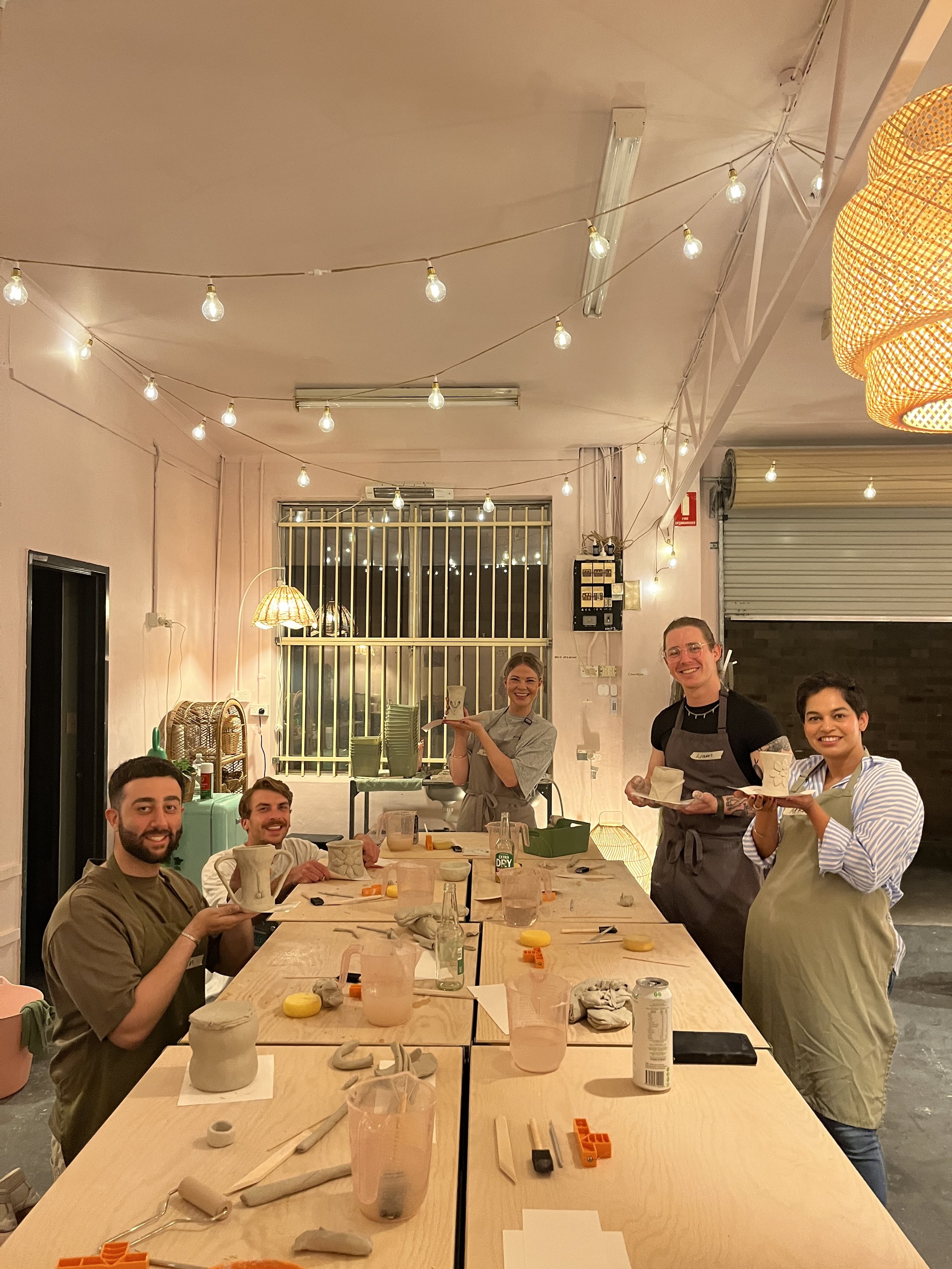 Group of people in pottery class holding clay creations, smiling at a table with tools and materials, under string lights in an art studio.