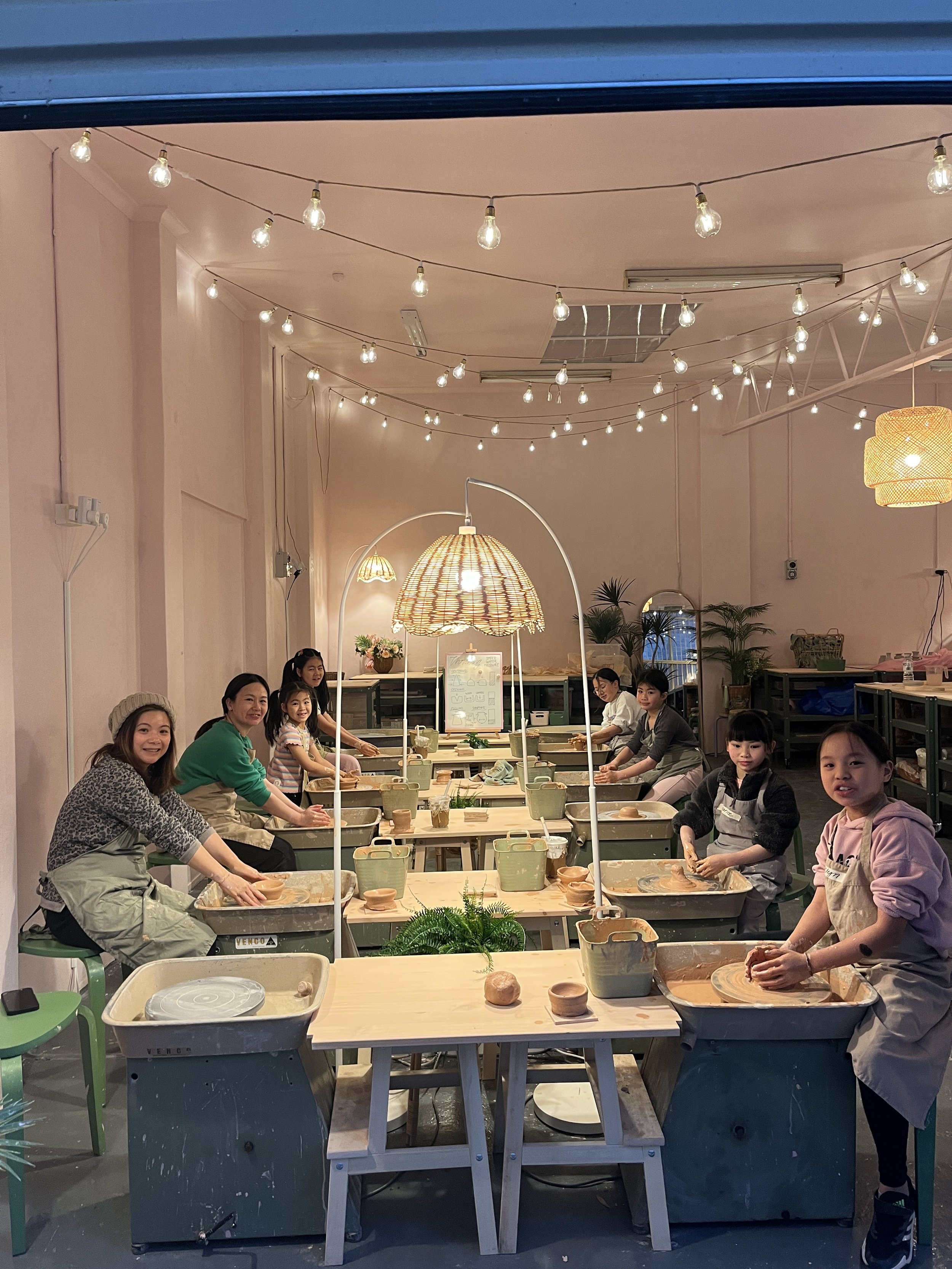 People in a pottery class at indoor studio with string lights.