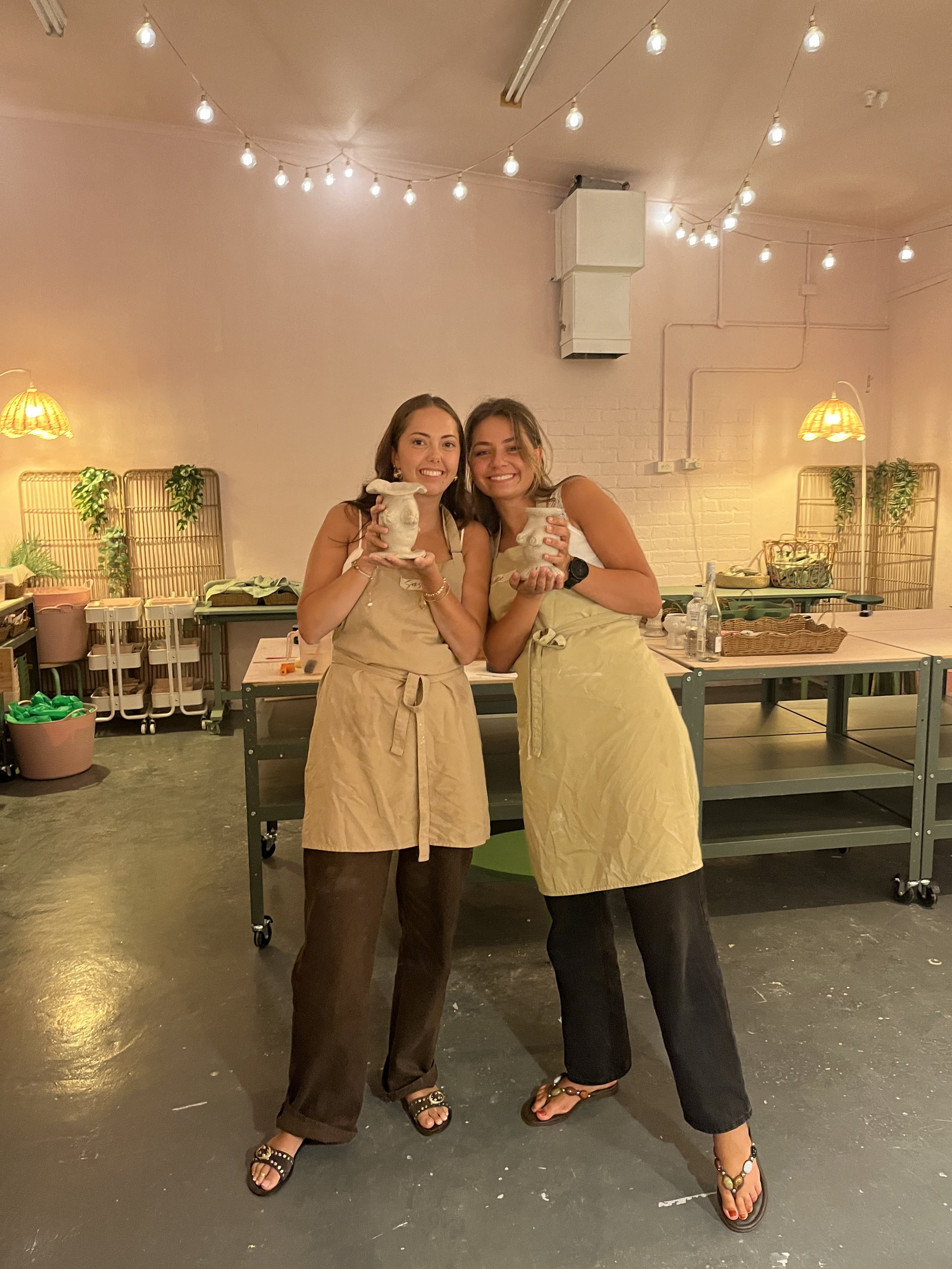 Two women in aprons holding clay pots in a pottery studio with string lights.