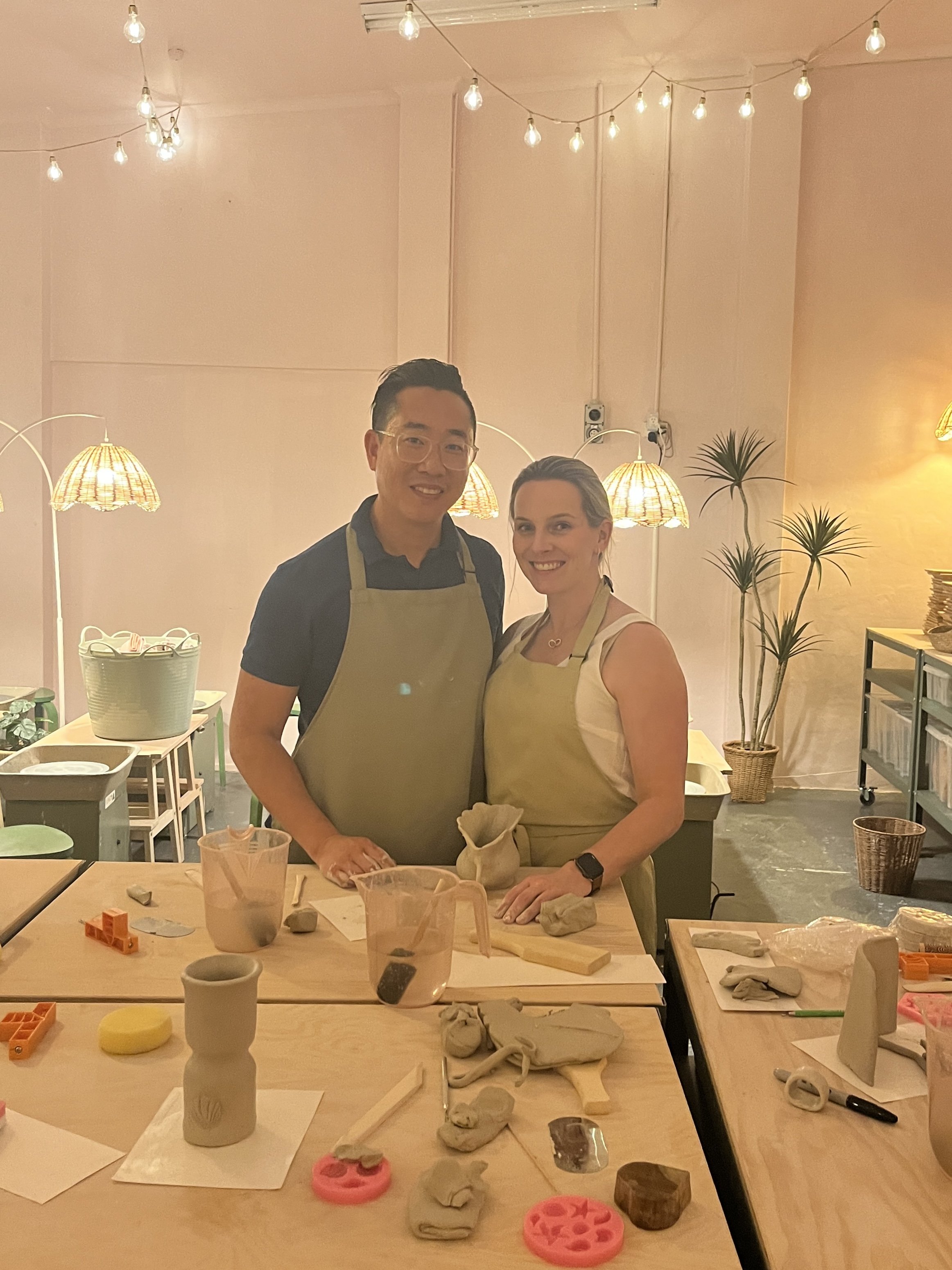 Two people wearing aprons standing in a pottery studio, surrounded by crafting materials and clay sculptures on a table, with soft ambient lighting and hanging lamps in the background.