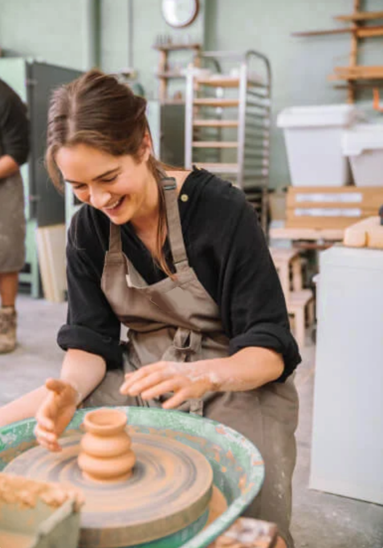 Person laughing while spinning clay on a pottery wheel in a workshop.