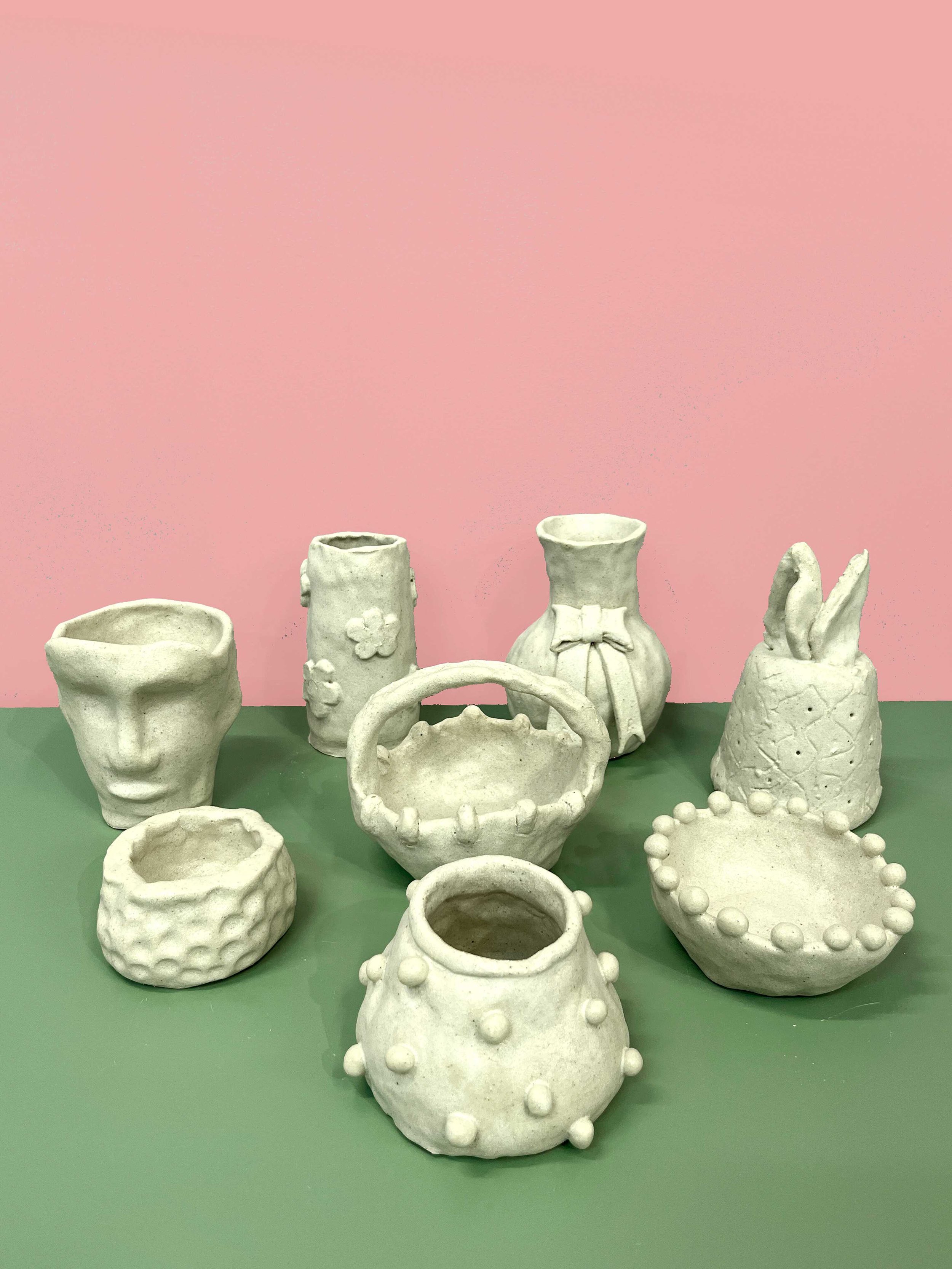 Handmade ceramic pots and vases with textured and decorative designs displayed on a green surface against a pink backdrop.