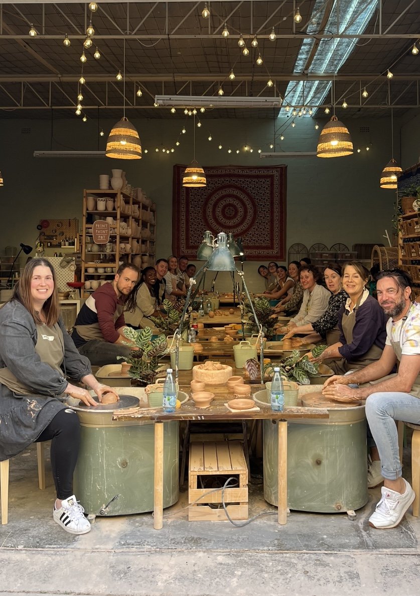 Group pottery class with people seated around tables, participating in pottery making, in a well-lit workshop with hanging lights and a decorative wall tapestry.