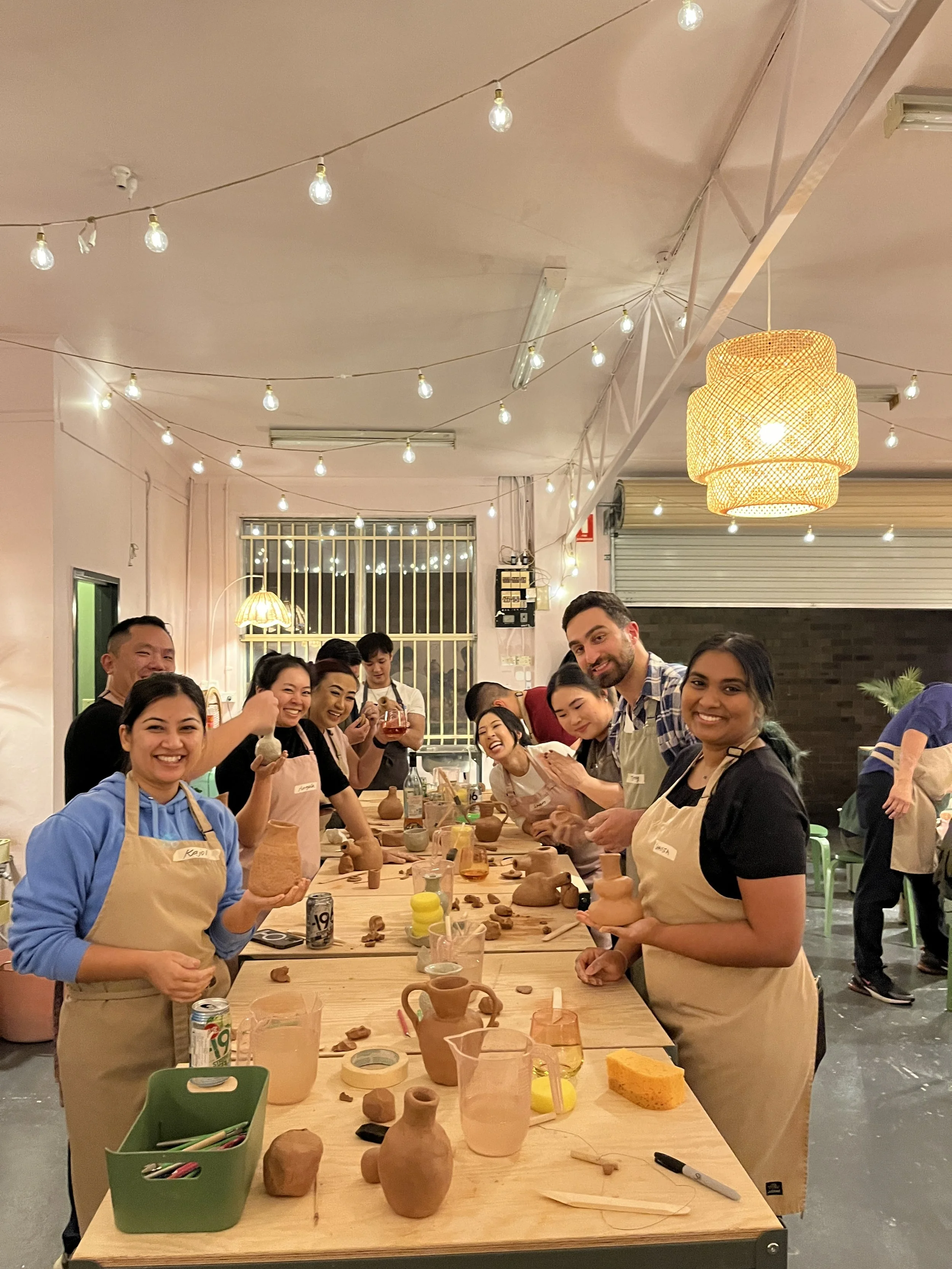Group of people participating in a pottery workshop at a long table filled with clay pottery, tools, and supplies, in a warmly lit room with string lights hanging from the ceiling.