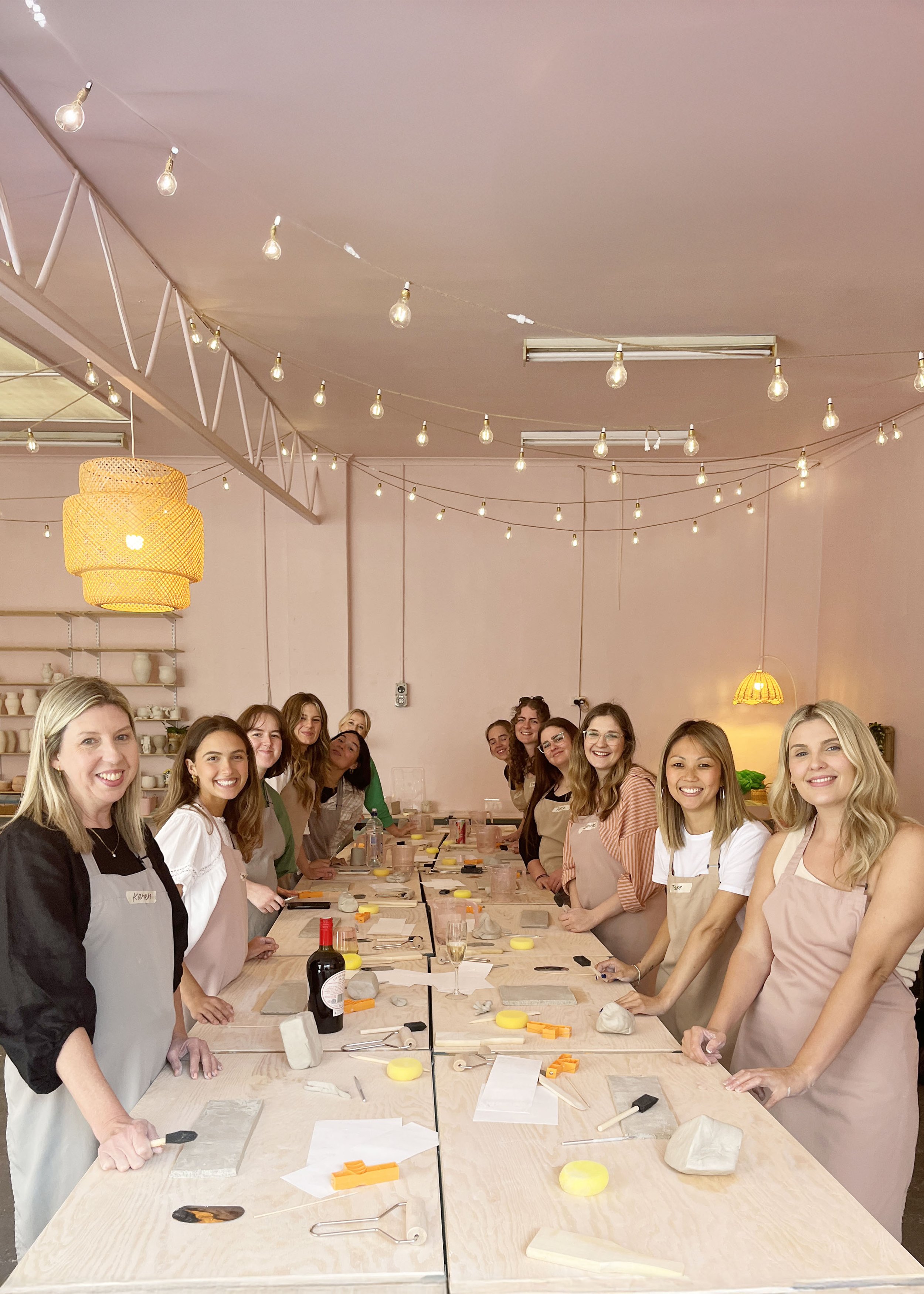 Group of women in a pottery workshop with clay and tools on tables, decorative lights overhead.