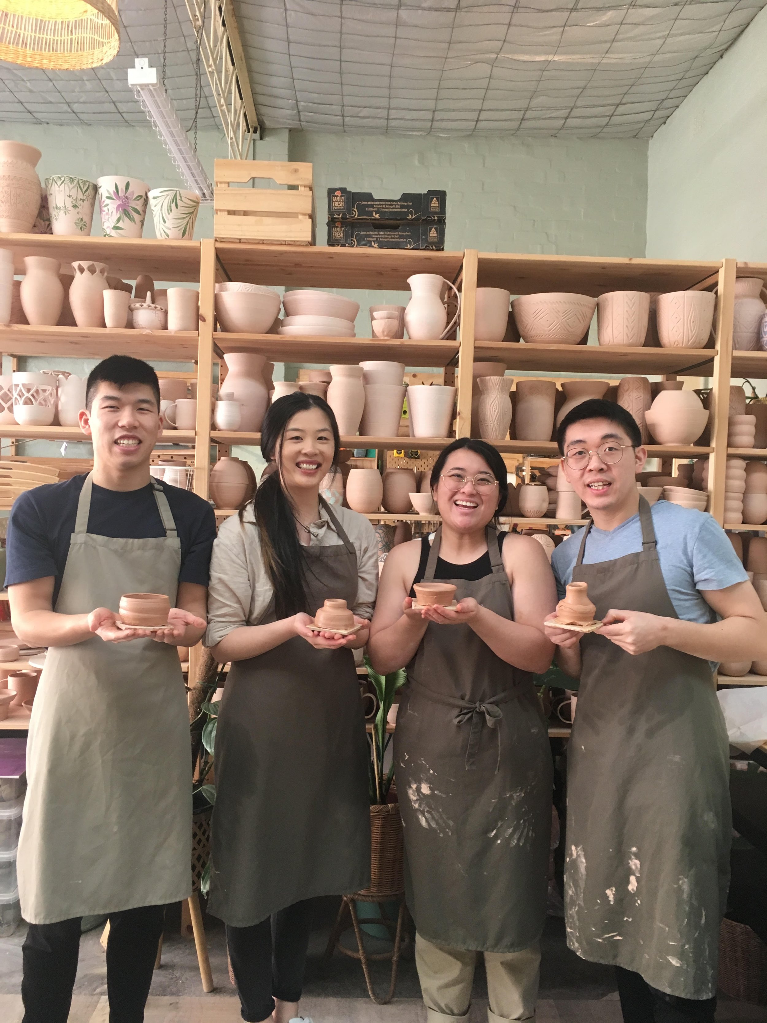 Four people in aprons holding pottery pieces in a workshop, with shelves of vases and pottery in the background.