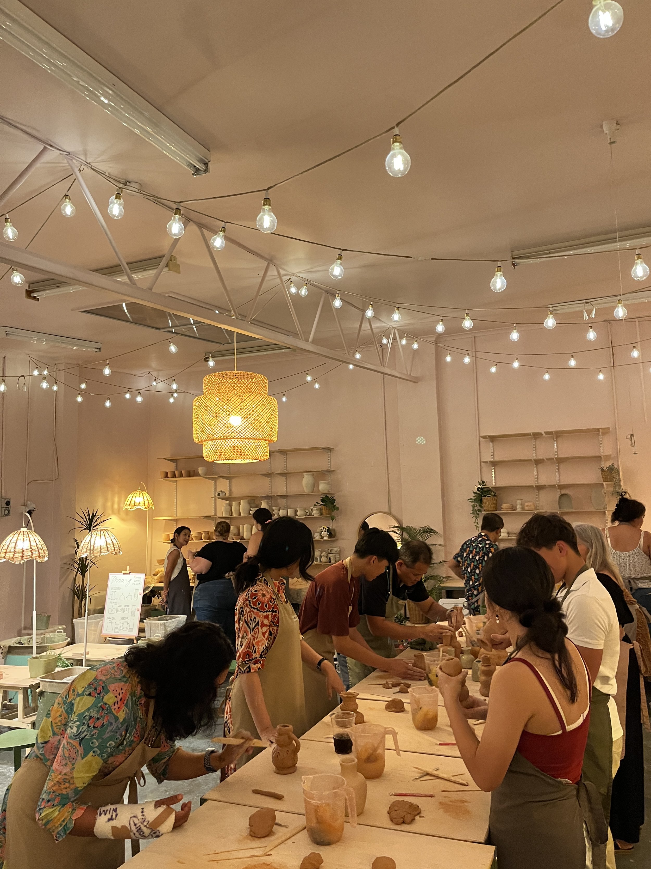 People in a pottery workshop creating clay sculptures in a well-lit studio with hanging string lights.