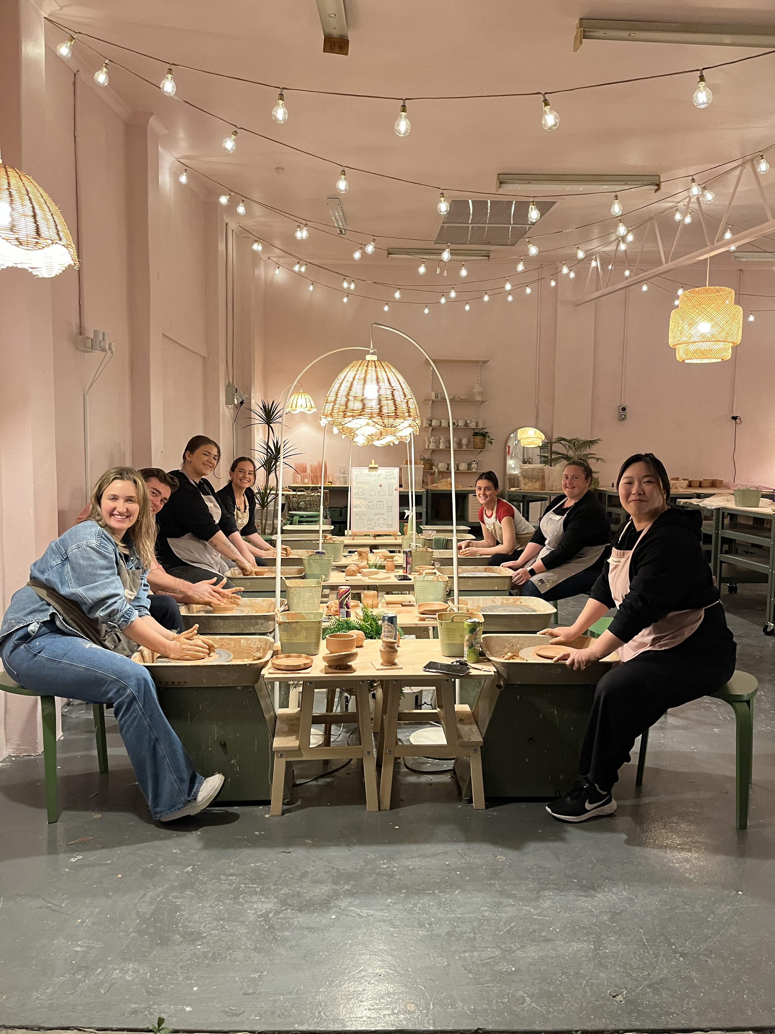 Group pottery class with participants seated at pottery wheels in a studio with decorative lighting and wooden shelves in the background.