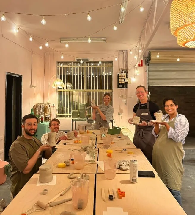 People in a cozy, decorated room celebrating at a long dining table with cups, plates, and art supplies, holding baked goods and smiling.