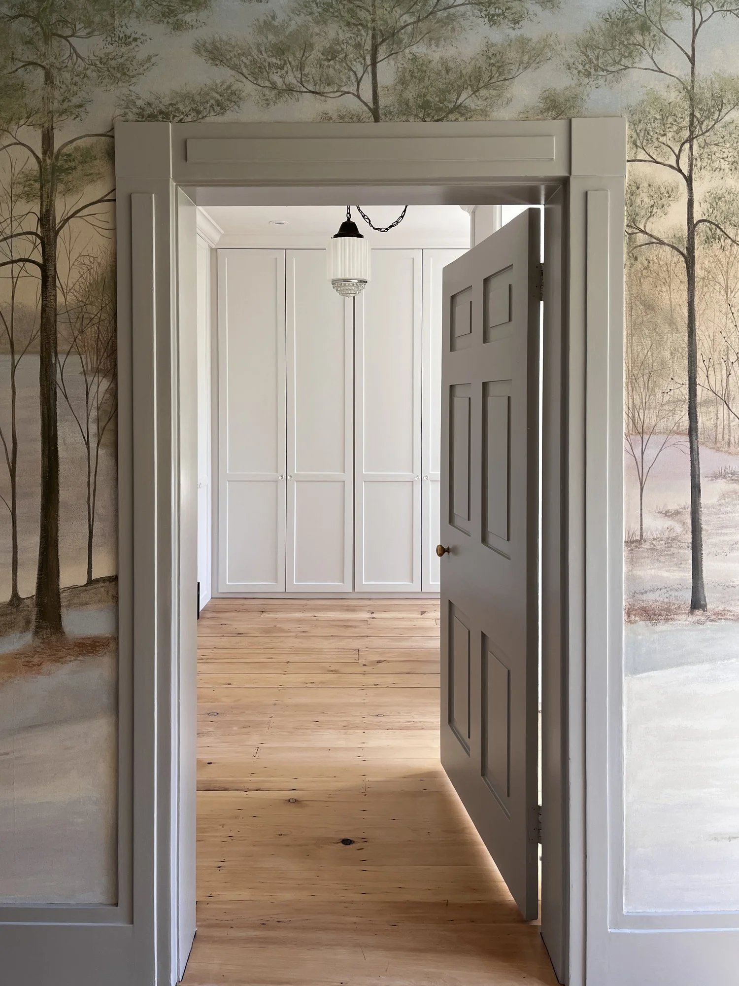 Room doorway with open six-panel door, white walls, wood floors, ceiling light fixture, and painted wall mural of trees and nature in the background.