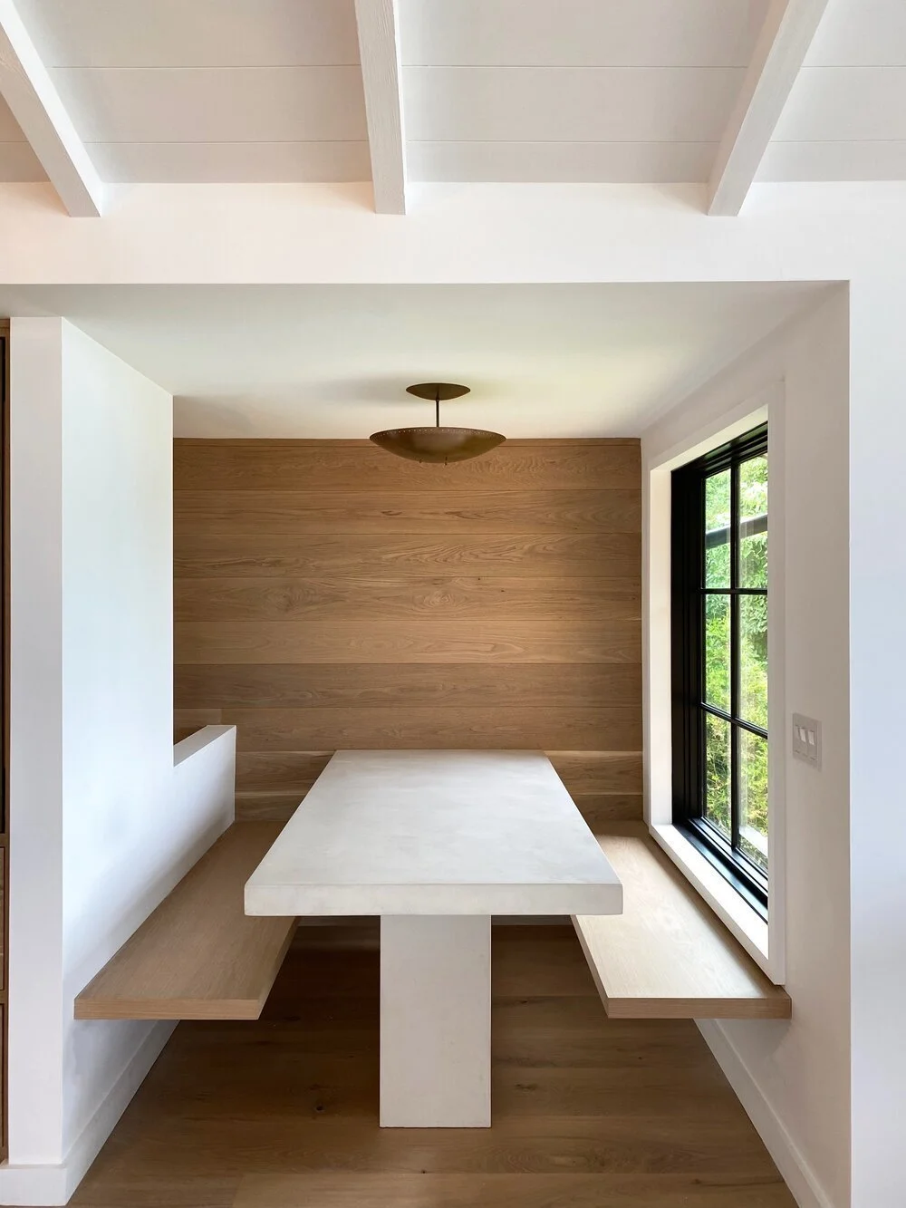 Interior dining nook with a built-in wooden bench seat, a white tabletop supported by a central pedestal, a wood-paneled wall, and a large window with black framing showing greenery outside.