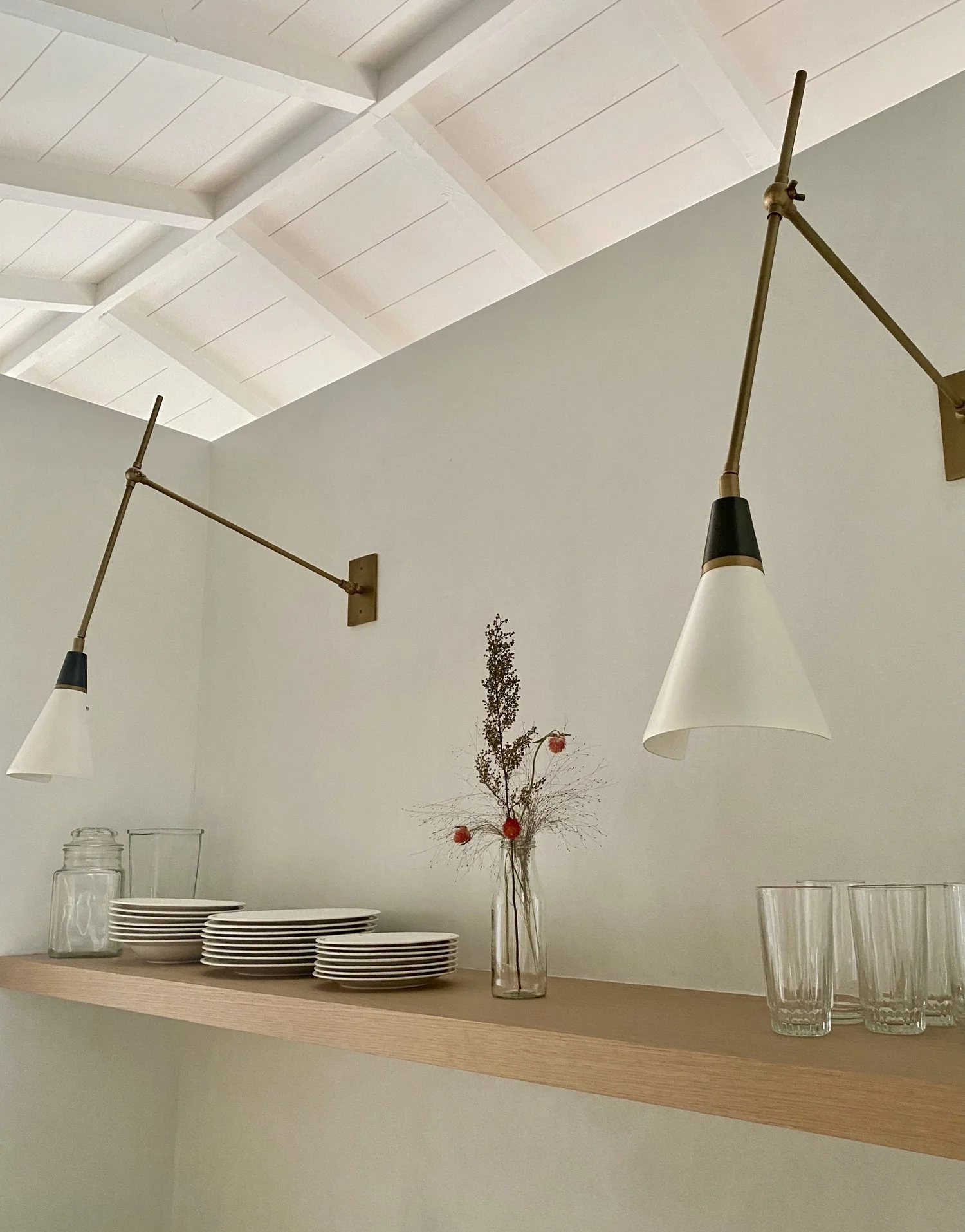 Wooden shelf with stacked white plates, a glass jar, a glass vase with dried flowers, and four drinking glasses, under white wall-mounted lamps with angled conical shades, against a white wall and ceiling with exposed beams.