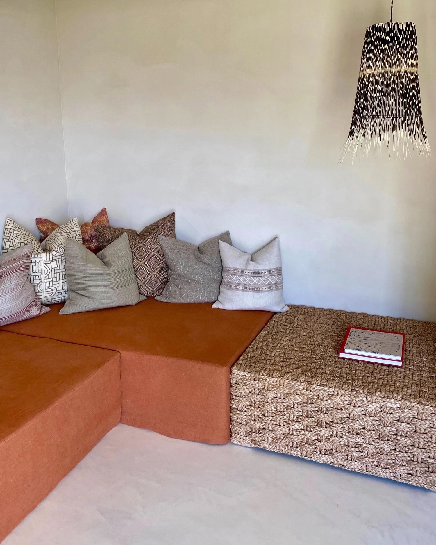 Corner of a living room with a sectional sofa covered in orange fabric, decorated with multiple patterned throw pillows, and a woven ottoman with a red book on top. A black and white woven pendant light hangs from the ceiling.
