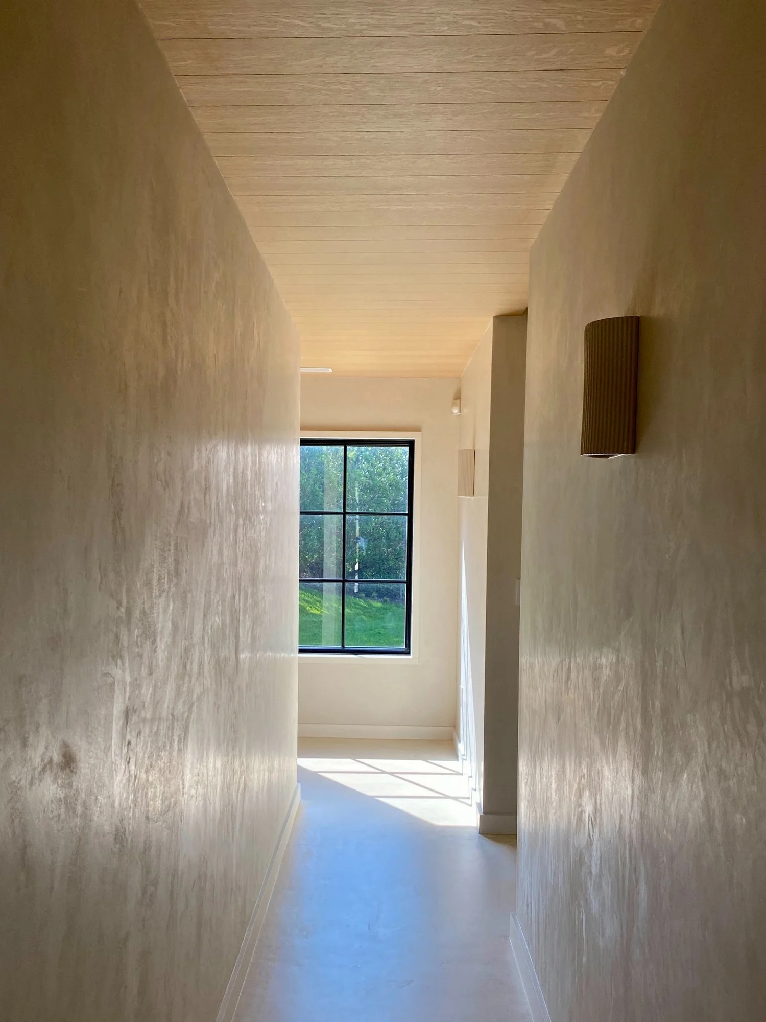 Empty hallway with beige walls, light-colored wooden ceiling, and a large window at the end showing green outdoor scenery.