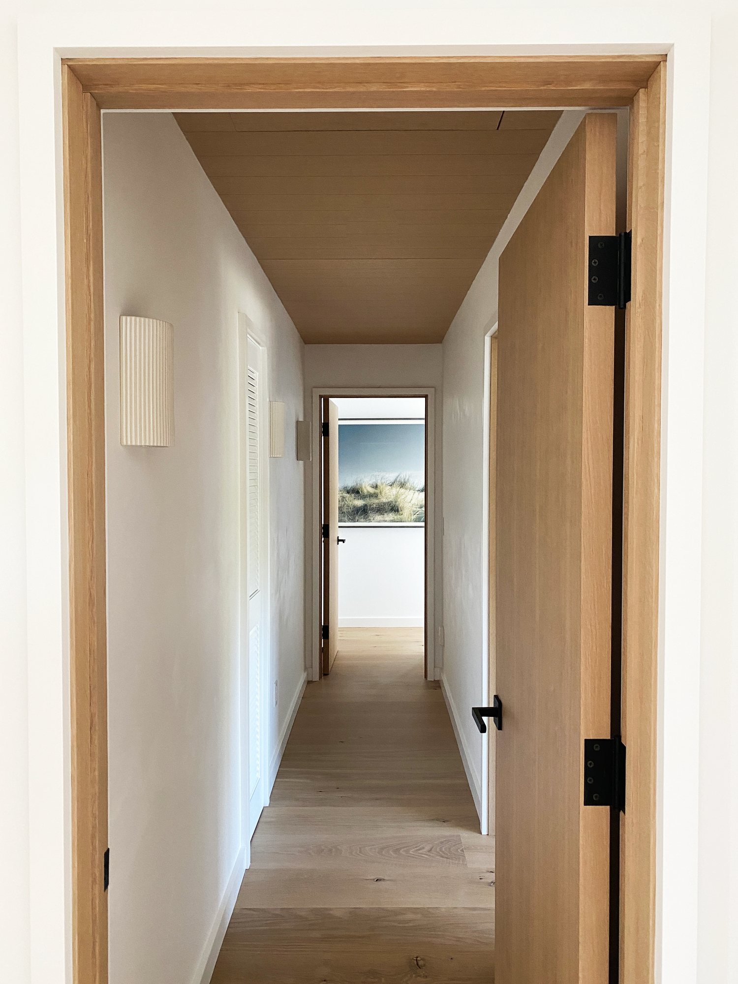 Interior view of a narrow hallway with light-colored wooden flooring, white walls, and a wooden ceiling, featuring a half-open door on the right and a door at the end showing a view outside with a field and sky.