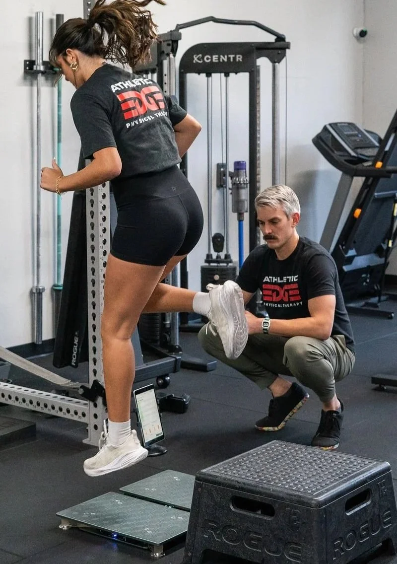A woman performing a leg exercise on a machine with a trainer assisting her by holding her foot in a gym.