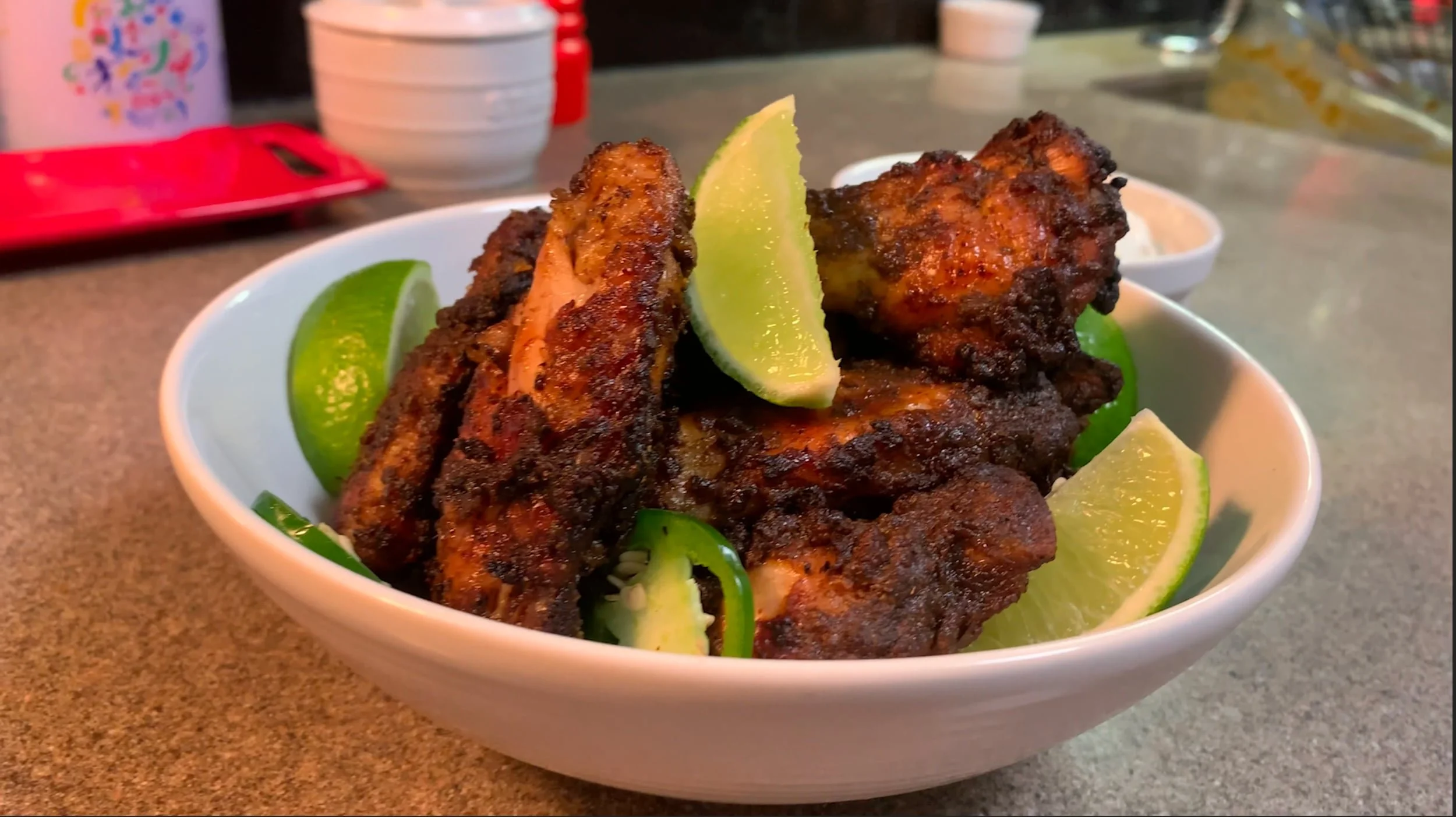 A bowl of air fryer jerk chicken wings garnished with lime wedges and sliced jalapeños on a kitchen counter.