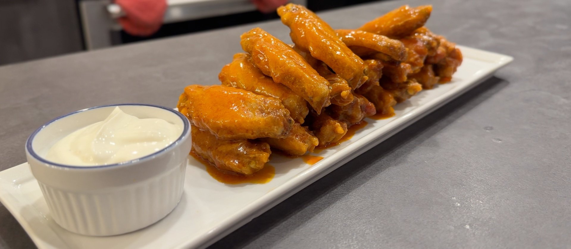 A plate of fried buffalo chicken wings served with a small cup of white dipping sauce on a gray countertop.