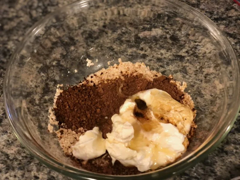 A glass bowl with espresso cheesecake overnight oats, cocoa powder, yogurt, and honey on a granite countertop.