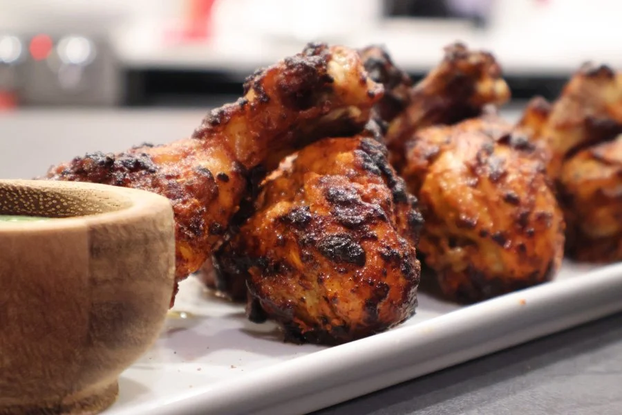 Close-up of air fryer tandoori chicken drumsticks on a white plate, with a wooden bowl of dipping sauce on the side.