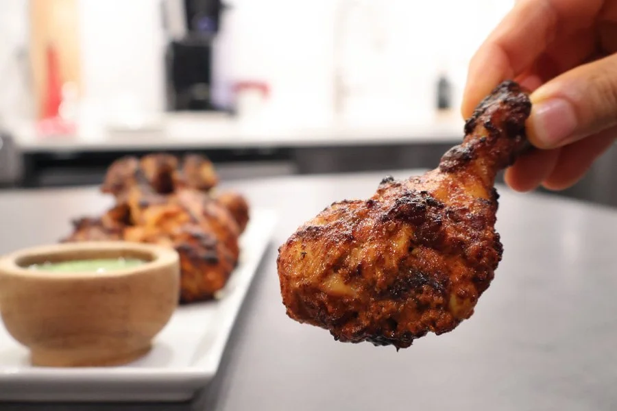 A hand holds a charred, grilled drumstick—like air fryer tandoori chicken—with more drumsticks and dipping sauce on a plate behind.