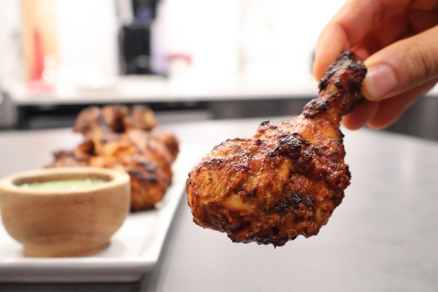 A hand holds a juicy air fryer tandoori chicken drumstick; more chicken and green sauce sit on a plate in the background.