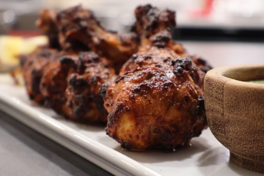 Close-up of air fryer tandoori chicken drumsticks on a white plate, served with a small wooden bowl of green sauce.