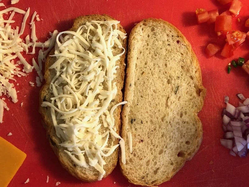 Two slices of bread, one with shredded cheese, on a red board—perfect for making an Indian grilled cheese sandwich with fresh veggies.