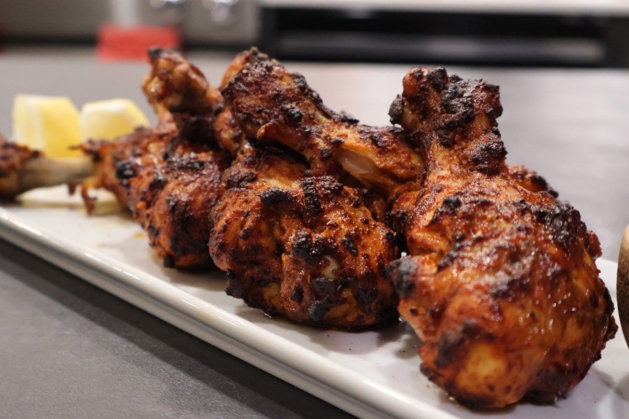 Four air fryer tandoori chicken drumsticks with char marks are served on a white plate, with lemon wedges in the background.