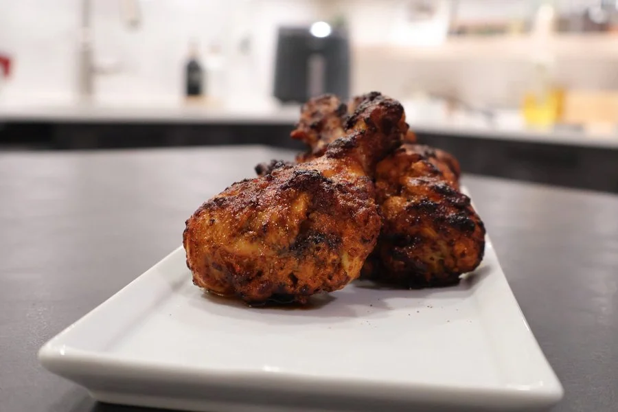 Three air fryer tandoori chicken drumsticks on a white plate, set on a gray countertop with a blurred kitchen background.