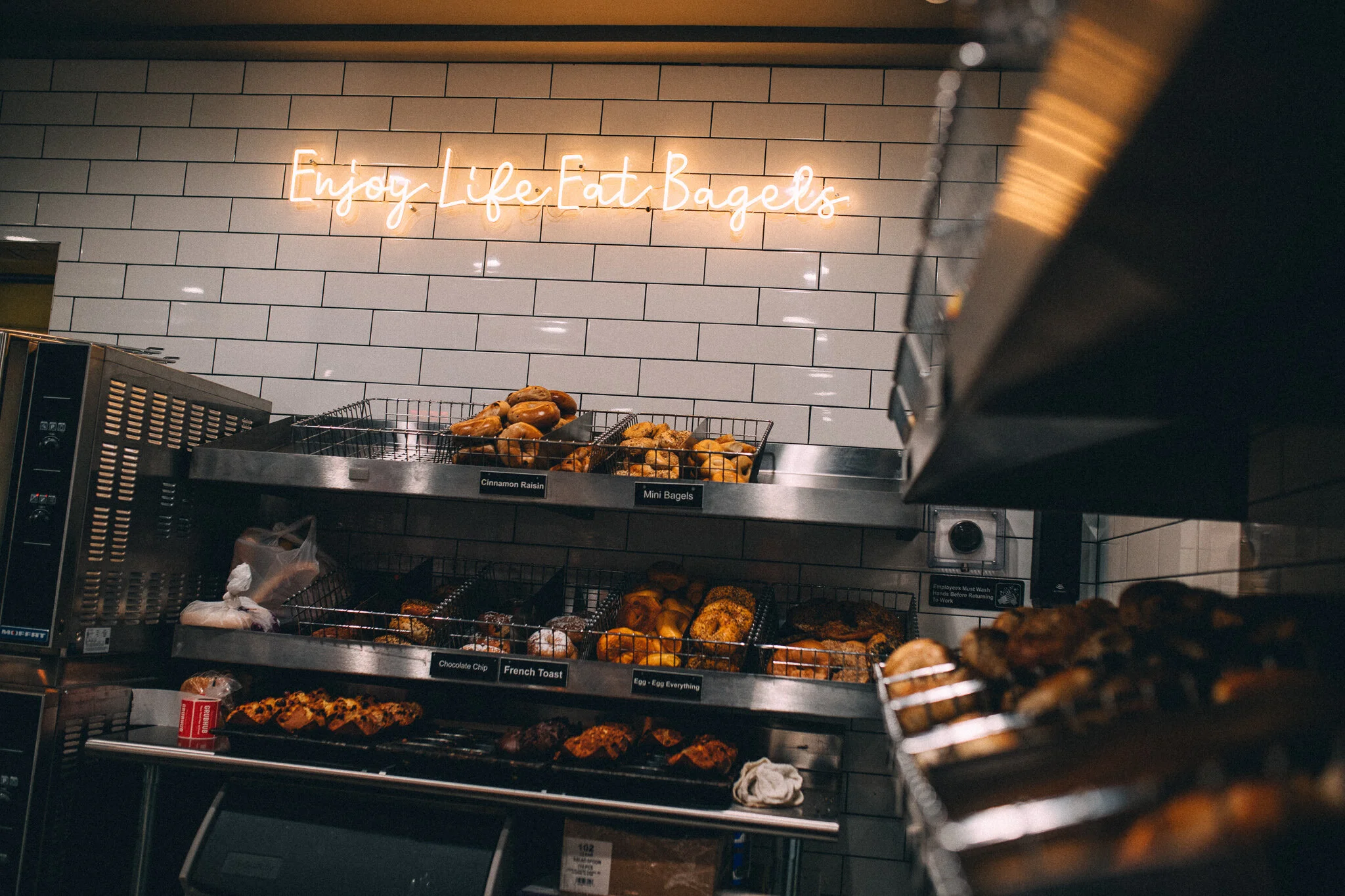 Enjoy Life Eat Bagels neon sign above the bakery racks inside Not Just Bagels