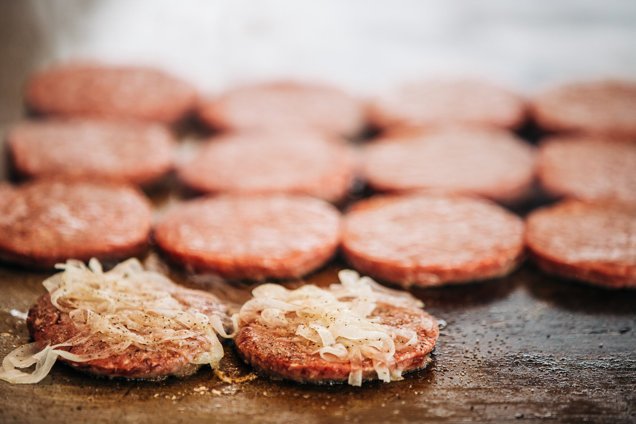 Rows of slider burger patties with sautéed onions cooking on the flat-top griddle