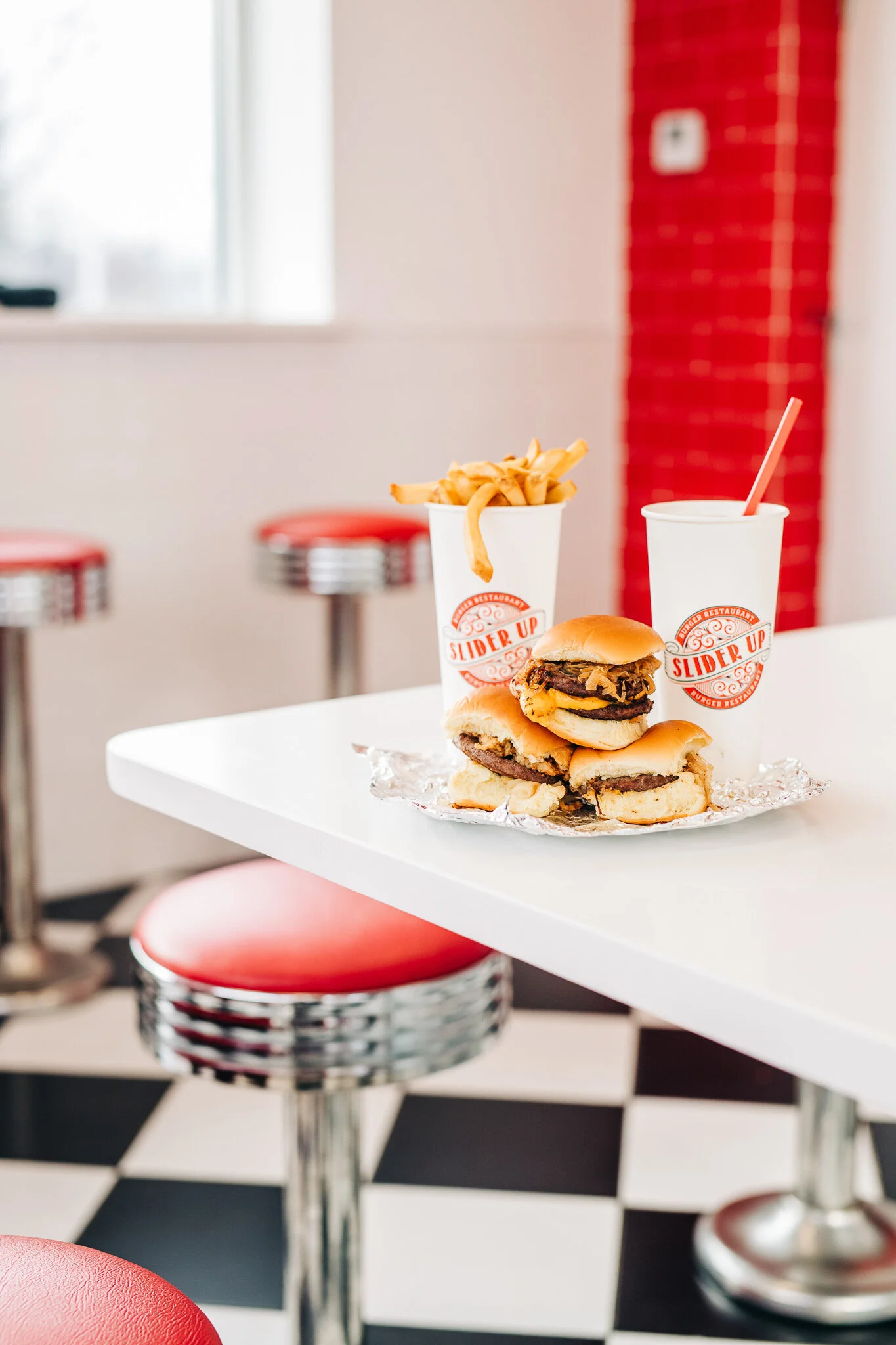 Stack of Slider Up burgers with branded fries and drink cups on a white diner table with red chrome stools