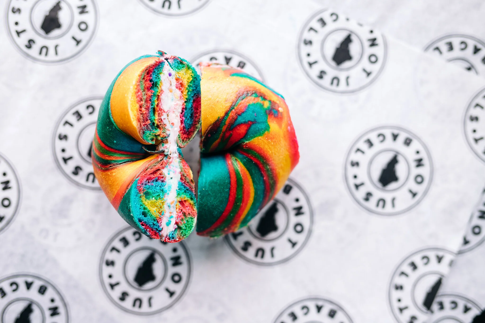 Sliced colorful rainbow bagel filled with cream cheese on Not Just Bagels branded paper