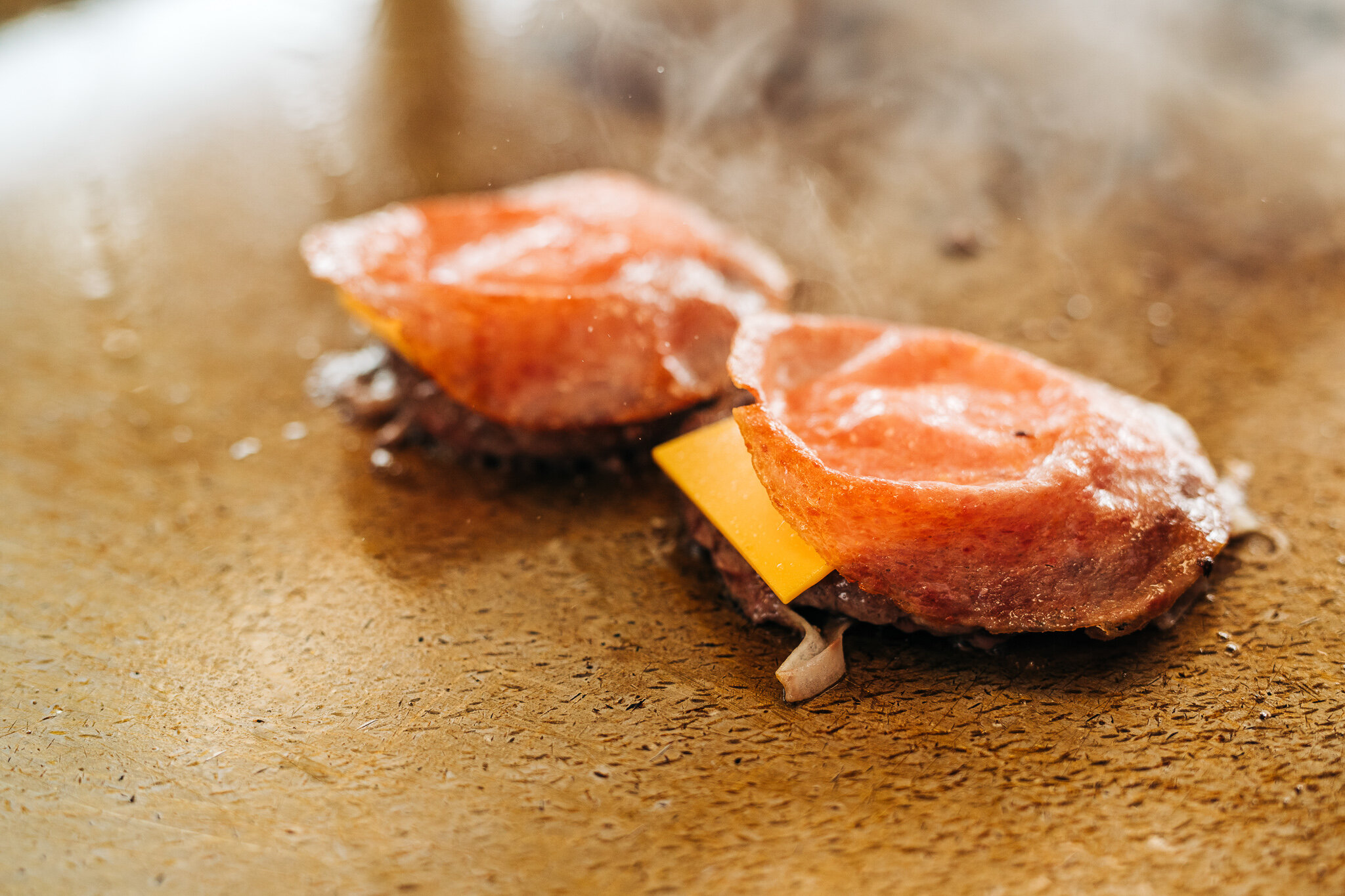 Two small burgers cooking on the flat-top with steam rising from freshly toasted buns