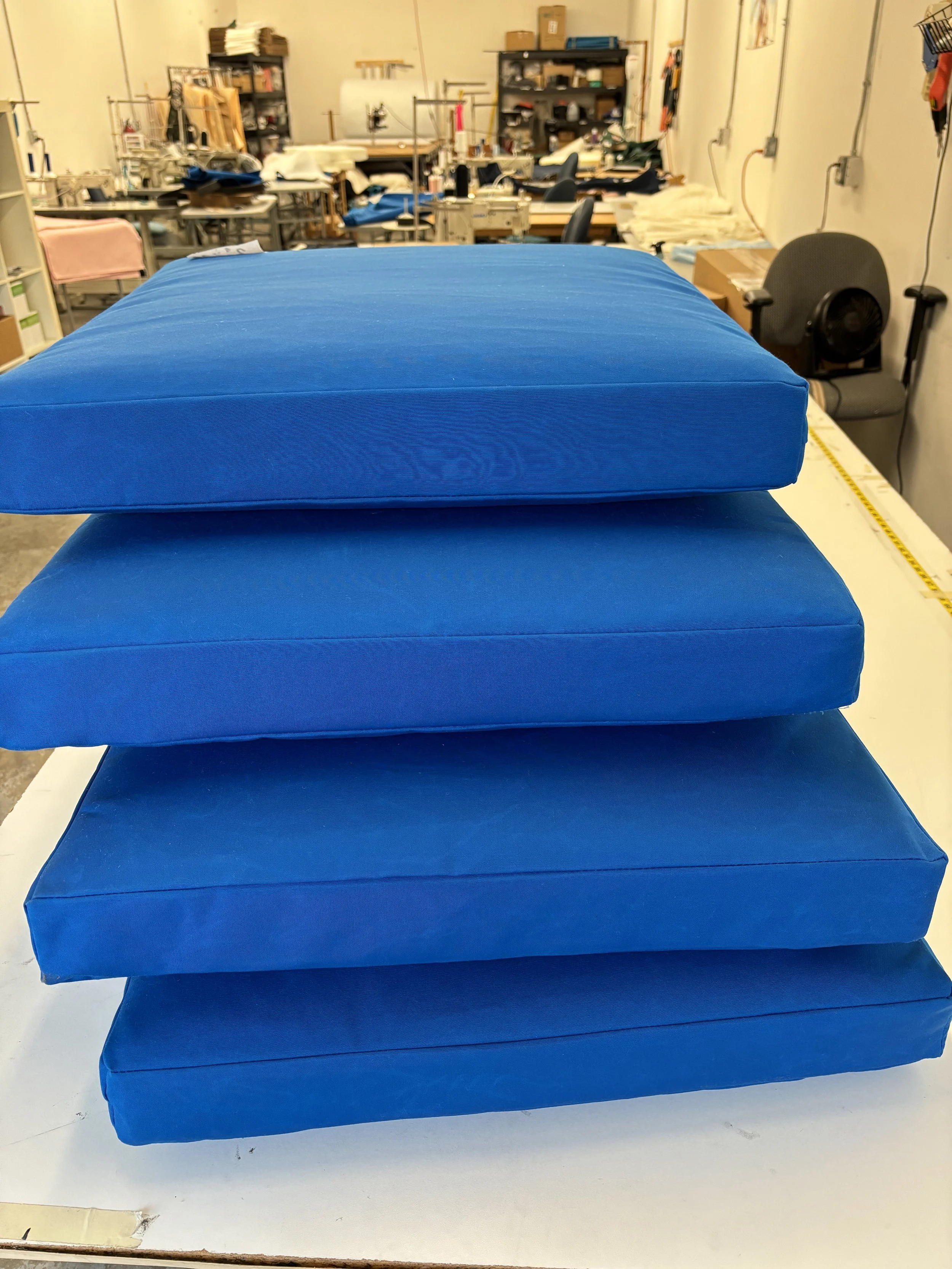 Close-up of three blue fabric cushions stacked on a white surface in a workspace filled with sewing machines, chairs, and shelves in the background.