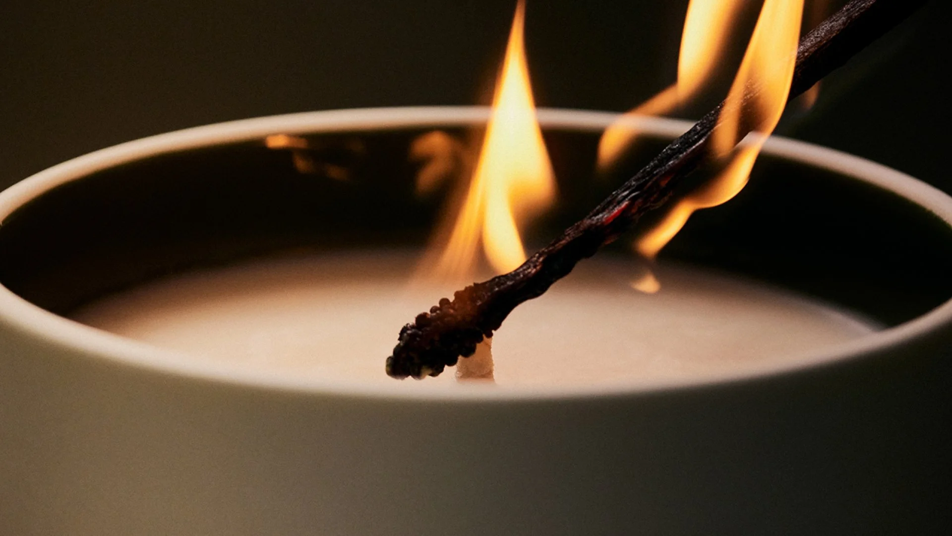 Close-up of a burning white candle with a dark wick, with a matchstick lighting the wick, producing flames.