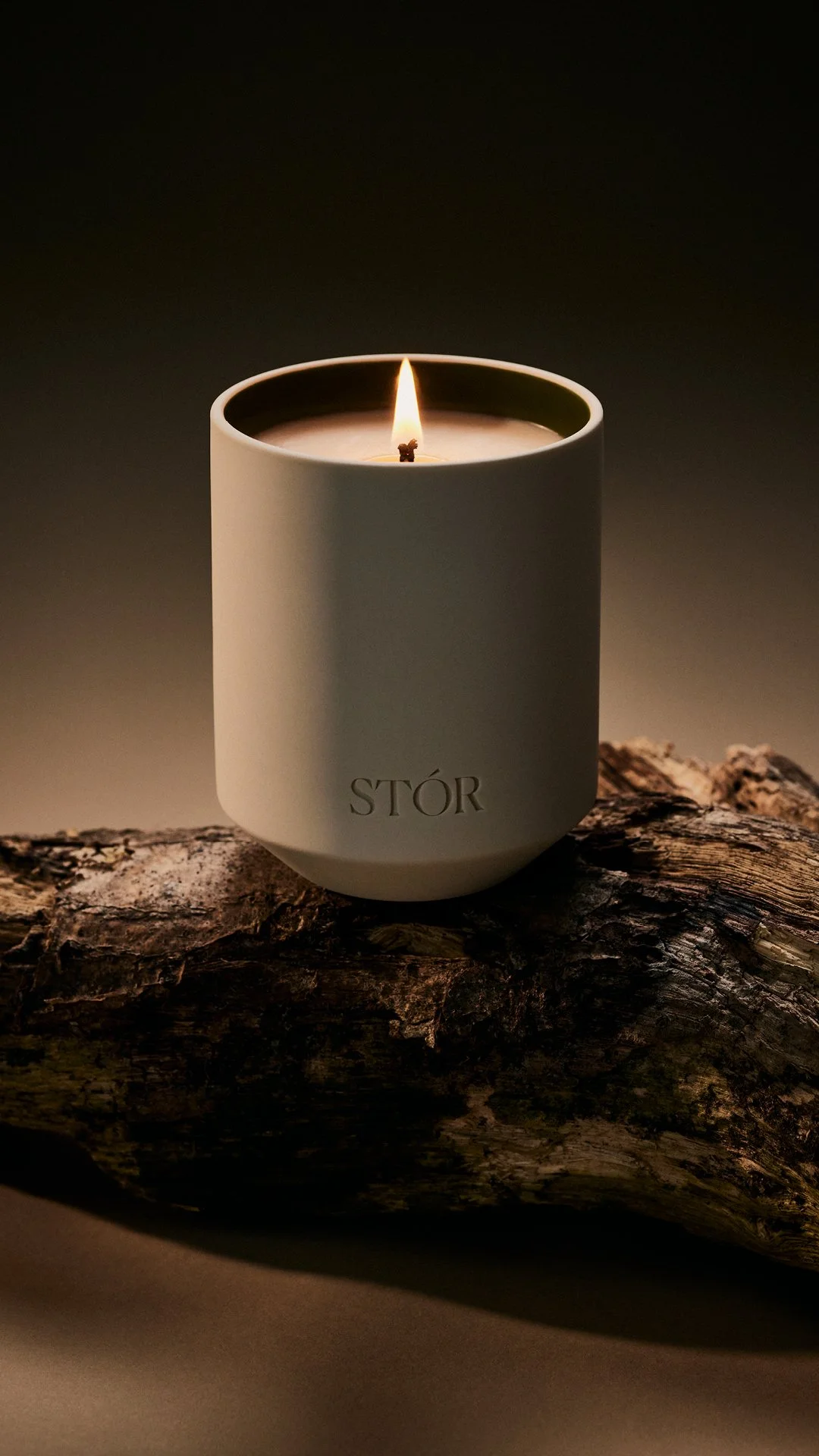 A white candle with the brand name 'STOR' in black letters, lit with a small flame, placed on a piece of textured wood against a dark background.