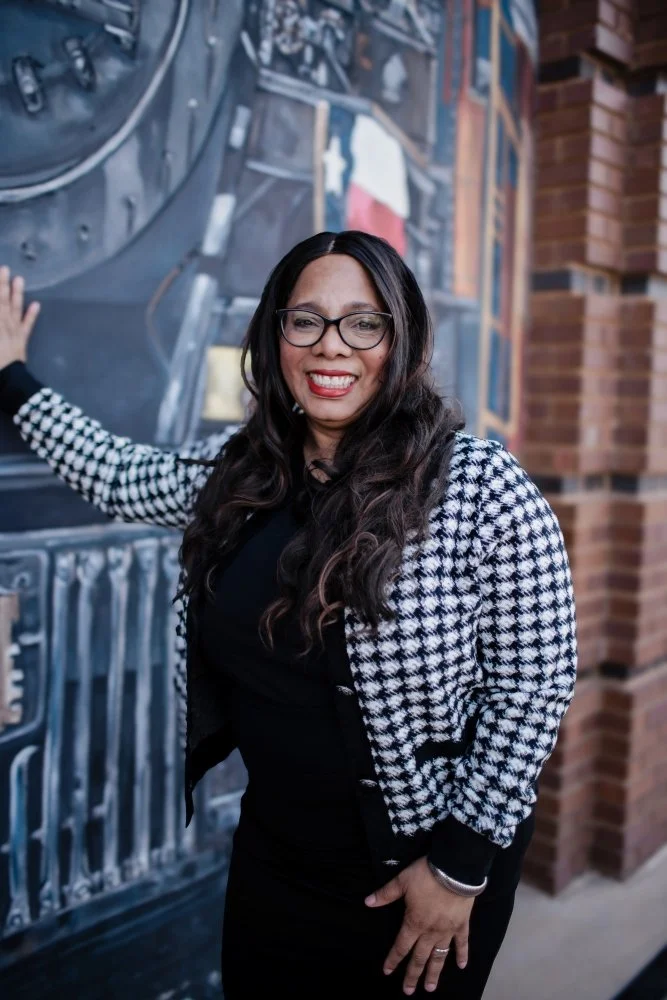 A woman with long dark hair, glasses, and red lipstick smiling, wearing a black dress and a black-and-white houndstooth patterned blazer, standing in front of a mural and brick wall.