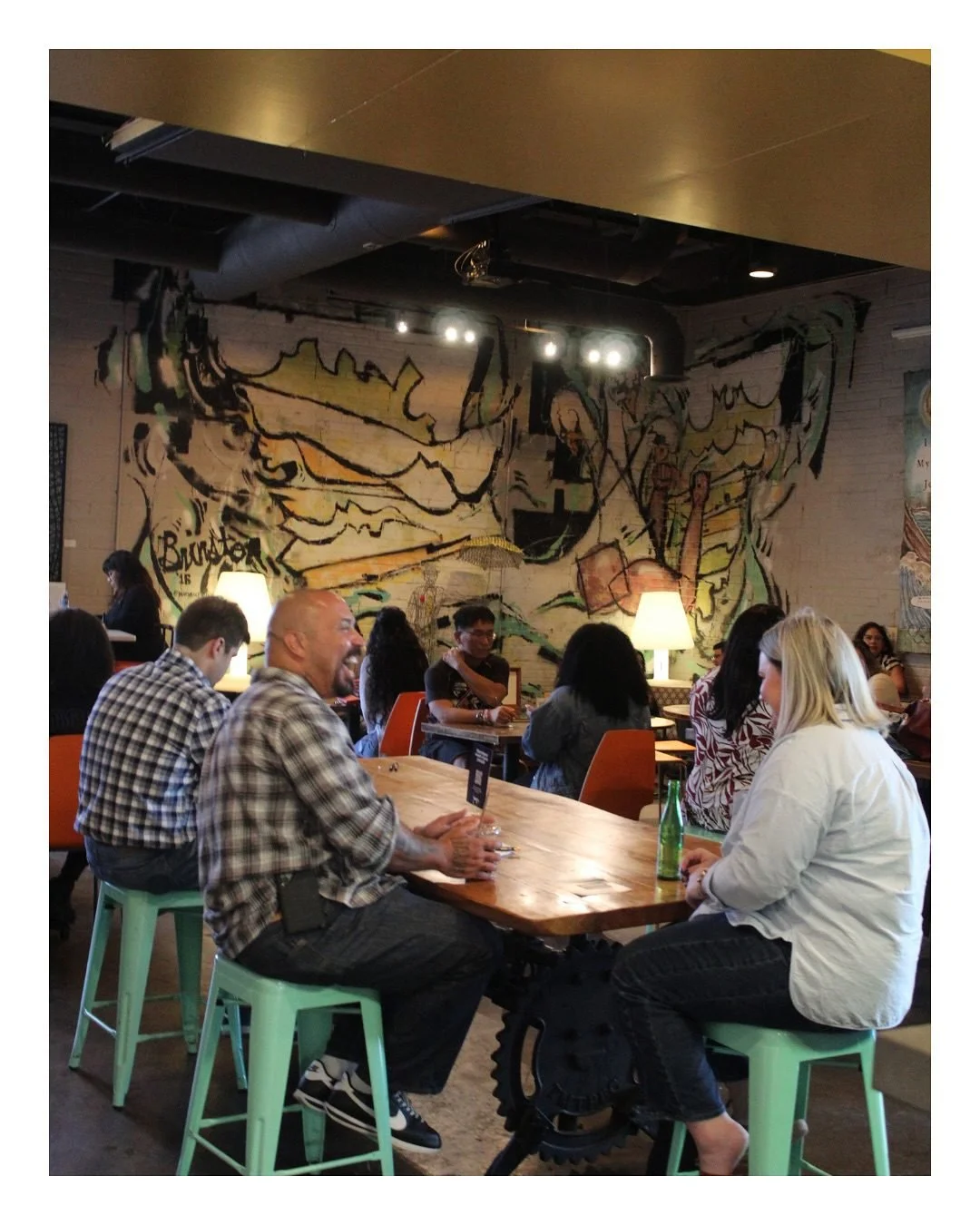 A lively cafe with people sitting around tables, some laughing and talking. The background features a large abstract mural on the wall and warm lighting.