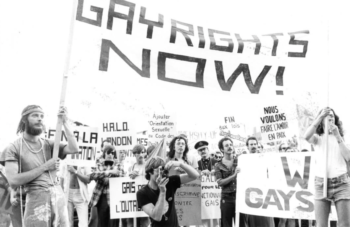 Black and white photo of a protest with people holding signs advocating for gay rights, including a large banner reading 'Gay Rights Now!', The Arquives Toronto LGBTQ+