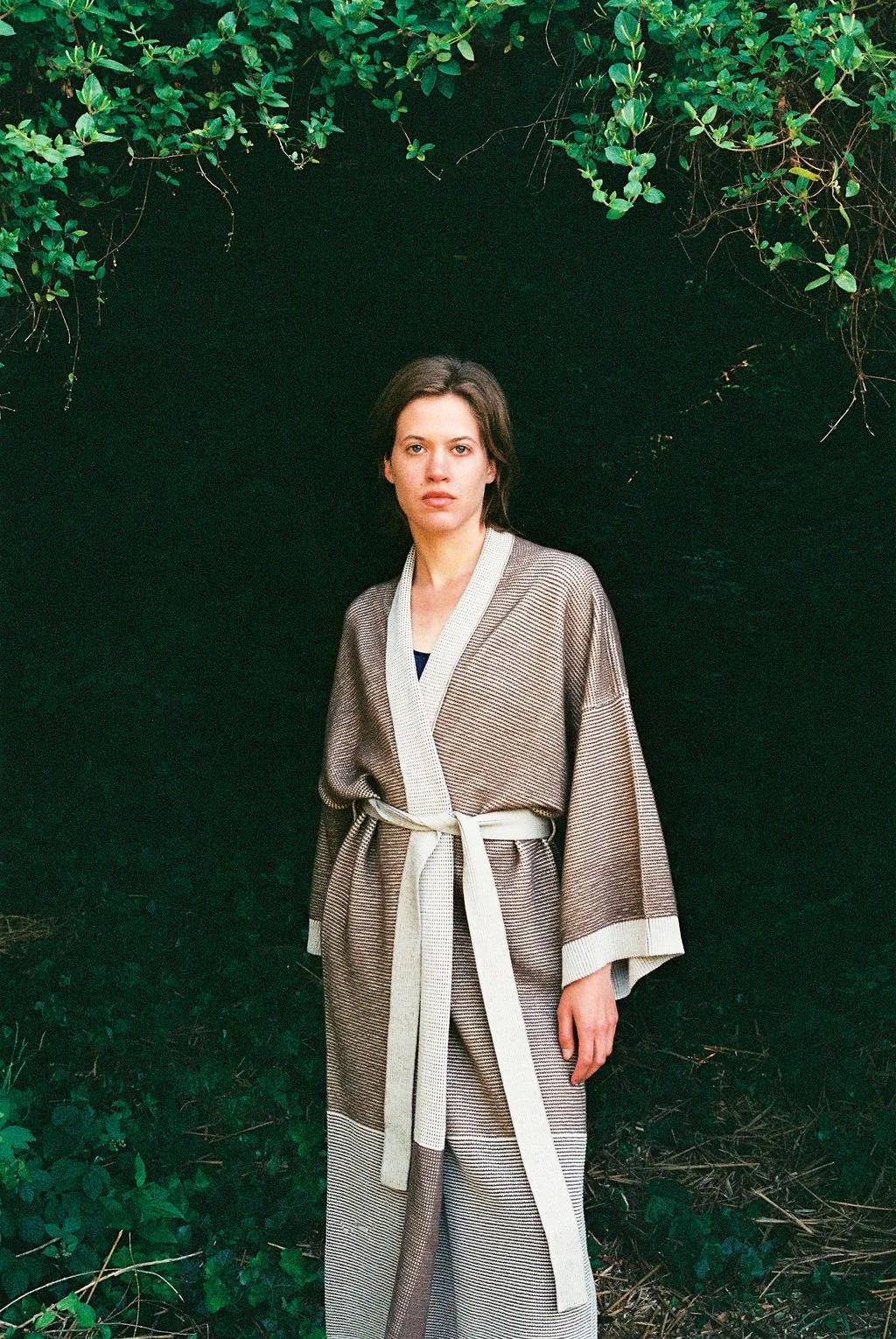 A woman standing outdoors in front of a dark green leafy background, wearing a beige and brown and beige knitted kimono with a belt.