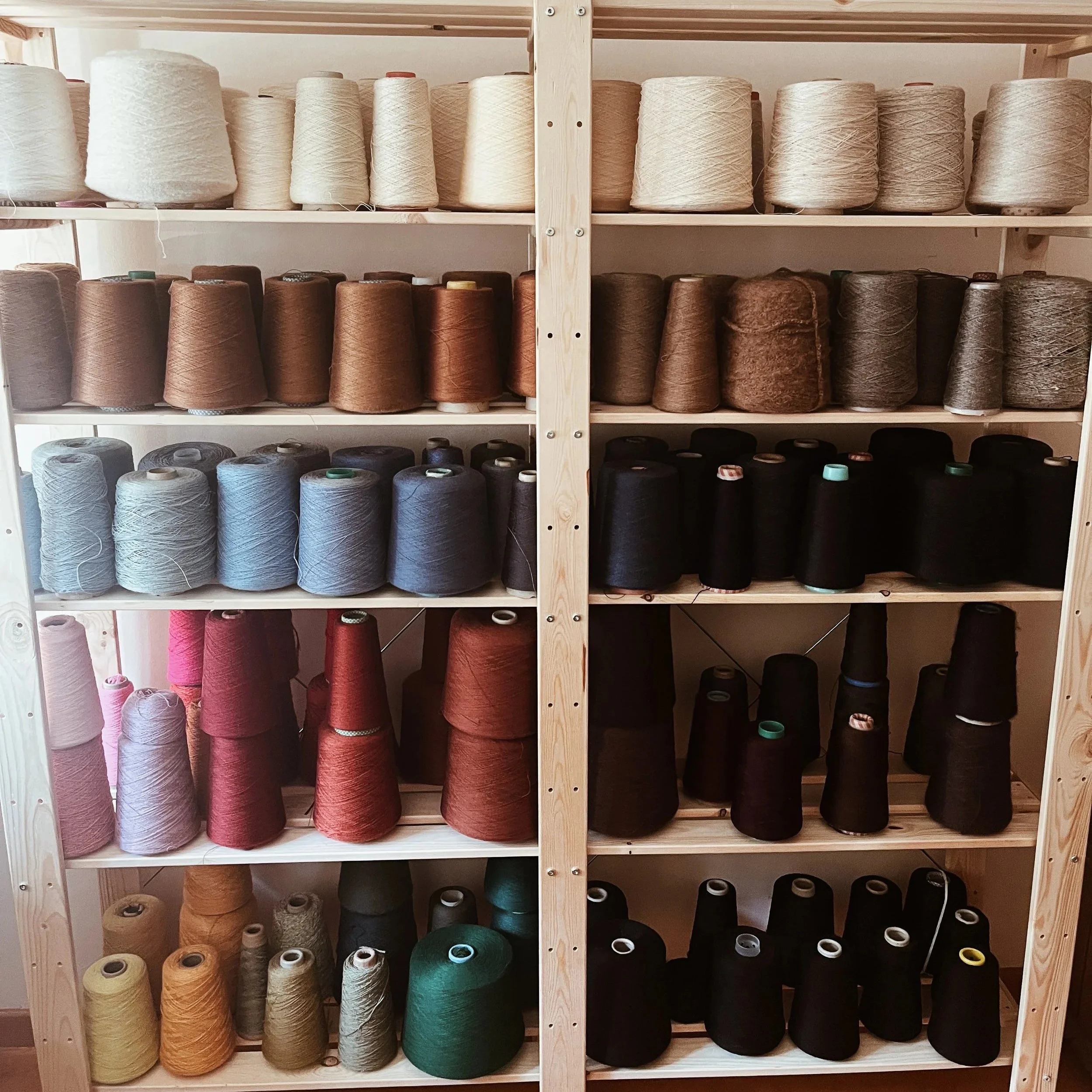 Wooden shelves filled with spools of deadstock yarn in various colors including white, brown, black, pink, orange, and green.