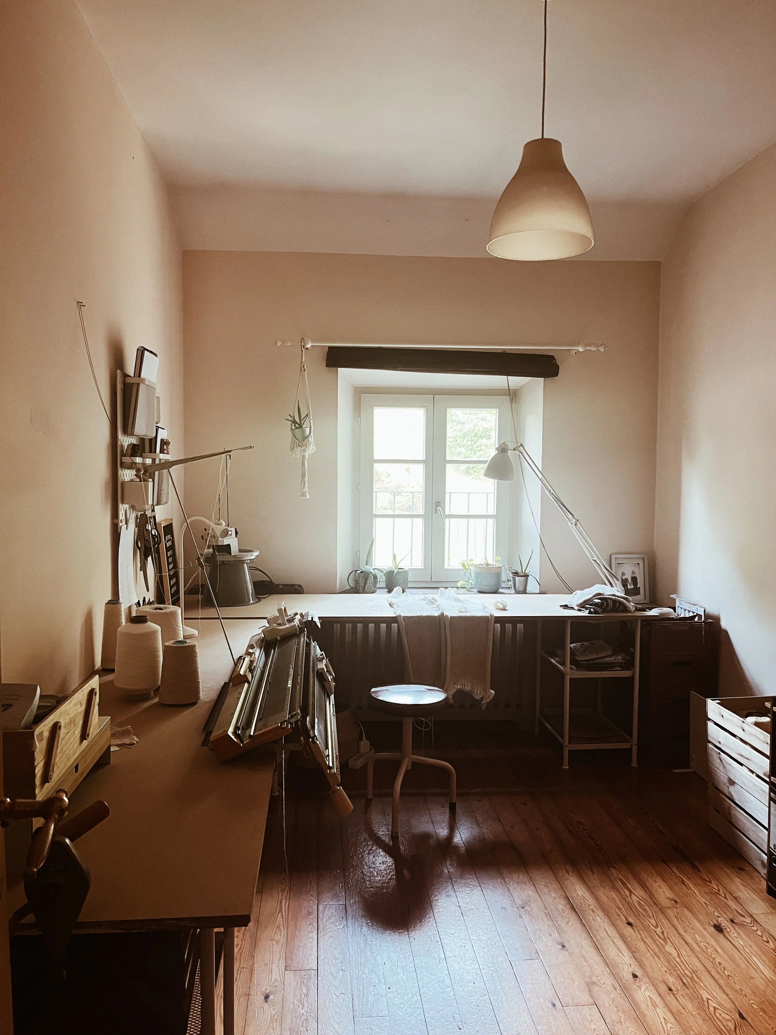 A home office with a window, desk, plants,  a knitting machine, and various items for crafting, knitting or sewing.