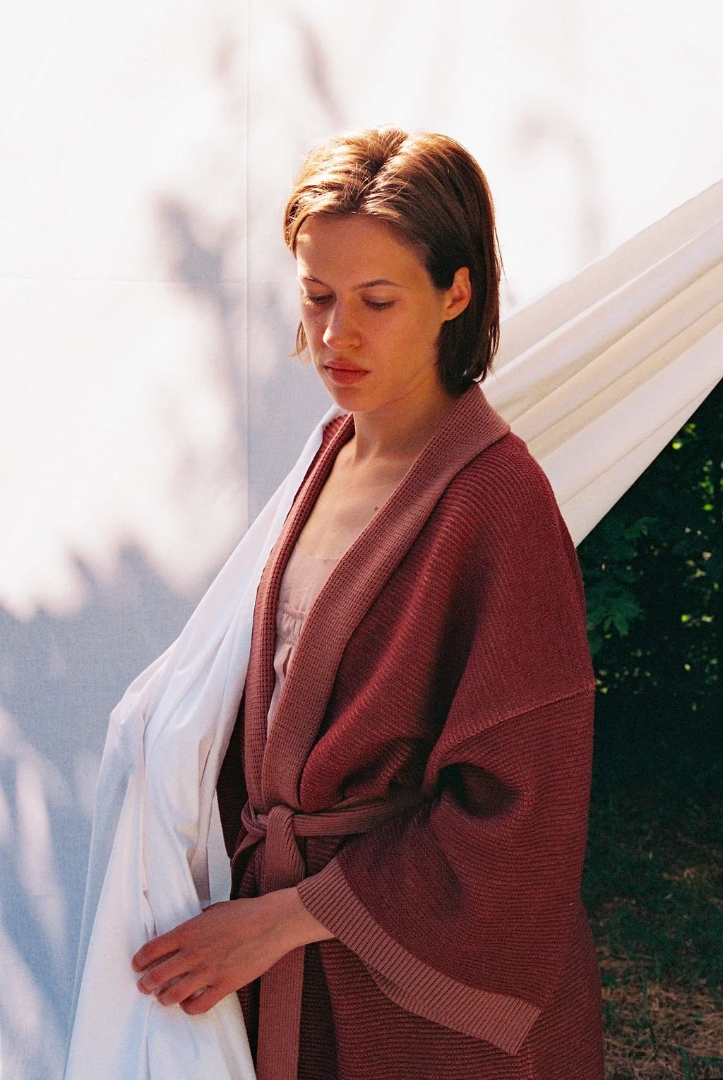 A woman with short brown hair, wearing a pink nightgown and a brick red knitted kimono coat, standing outdoors against a light-colored wall and green foliage, looking down with a contemplative expression.